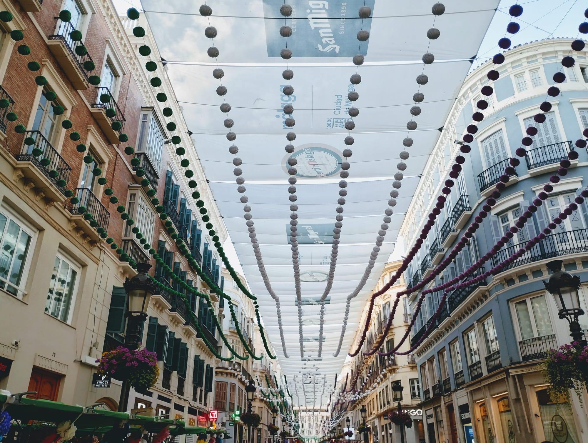 View of a pedestrian street with white canvas awnings and decorative beaded garlands suspended overhead, framed by colourful buildings.