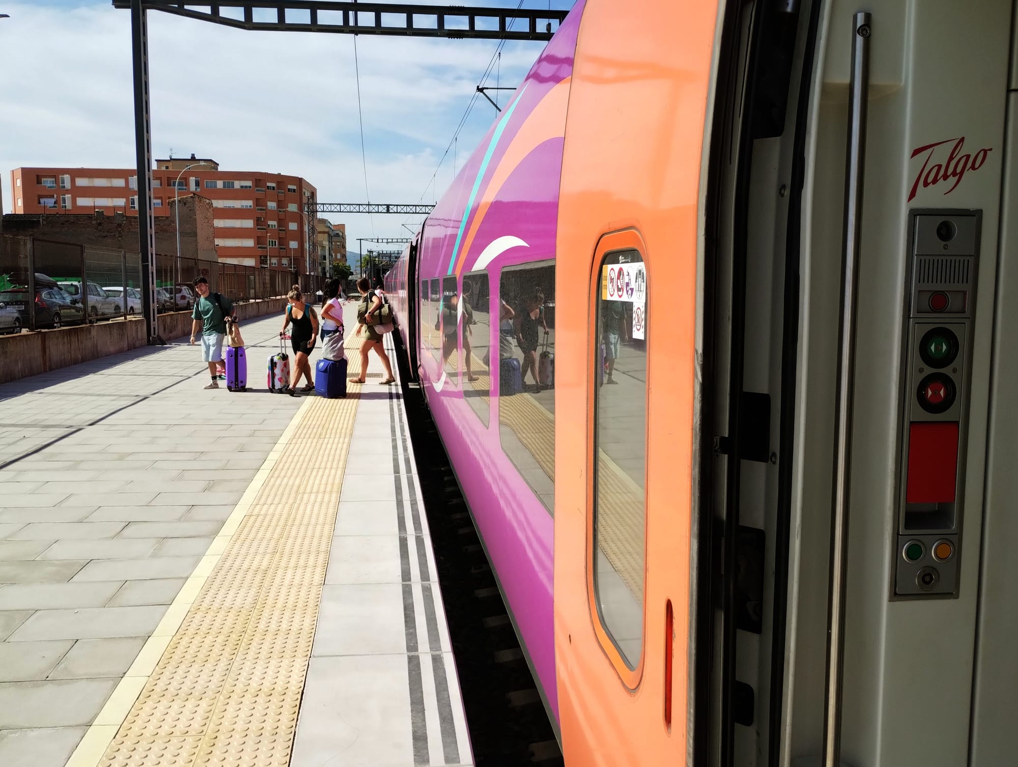 A vibrant pink and orange Talgo train at a station platform, with passengers awaiting the train.