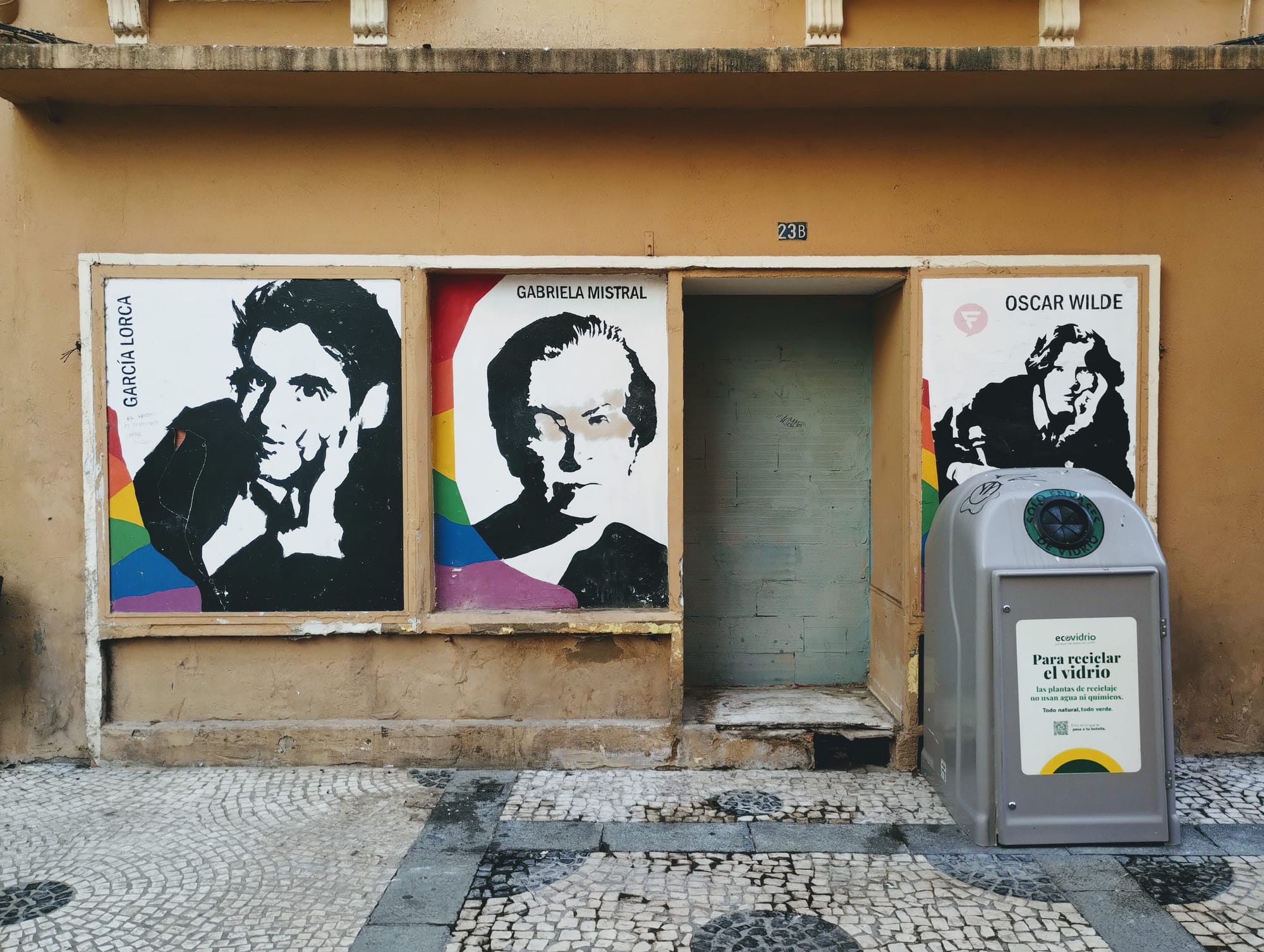 Exterior wall with three black-and-white portraits of literary figures: Garcia Lorca, Gabriela Mistral, and Oscar Wilde behind a glass recycling bin. The door to 23B is bricked-up with grey concrete blocks.