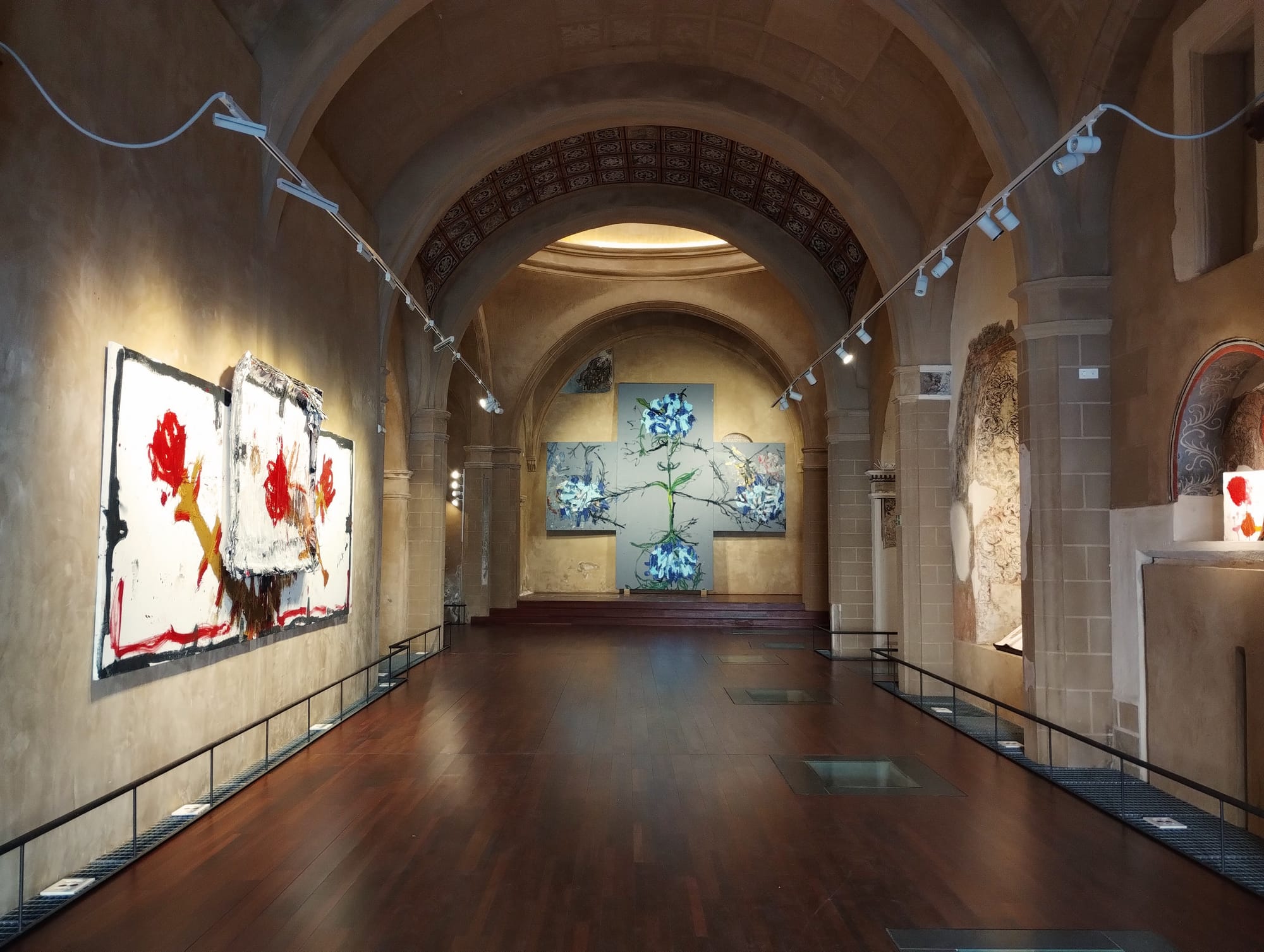 Gallery in a deconsecrated church with arched ceilings, dark wooden floors, and displayed abstract paintings, lit by track lighting, leading to a central floral painting installation.