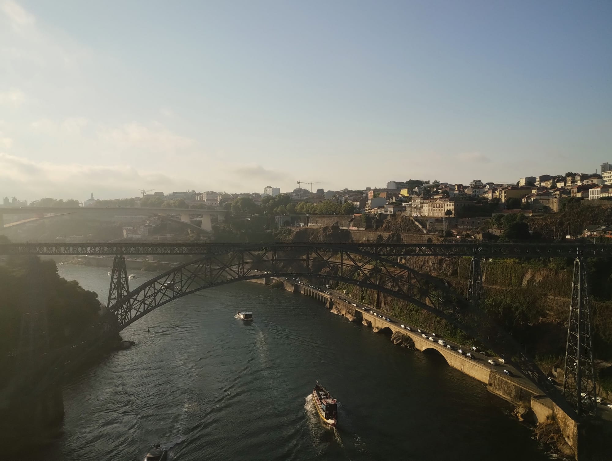 A view of the Dom Luís I Bridge spanning the Douro River, photographed from a train crossing an adjacent bridge. Boats sail along the river.