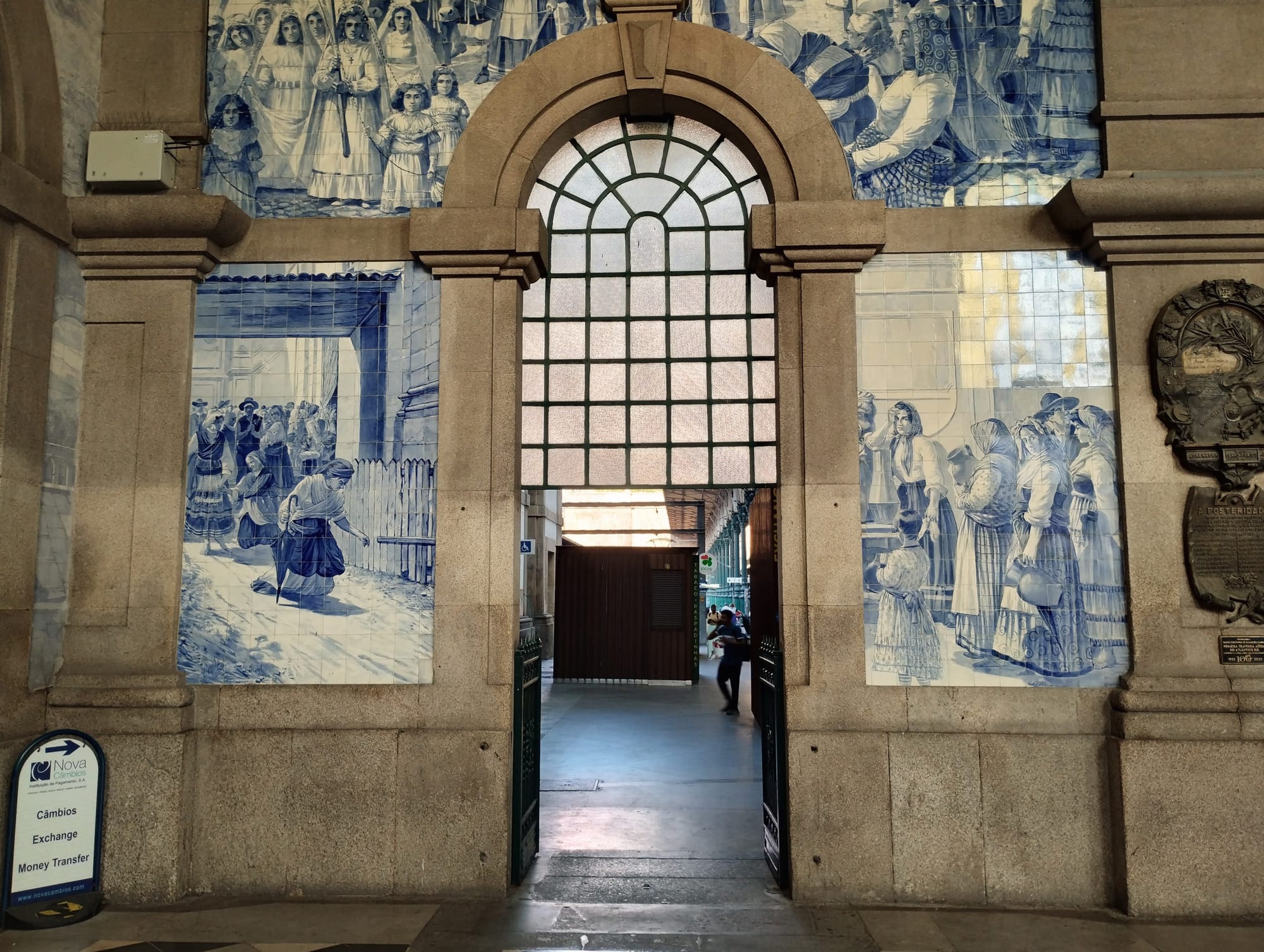 A door in Porto Sāo Bento station, with blue and white tiled murals depicting scenes of people, flanked by stone columns and an arched window.