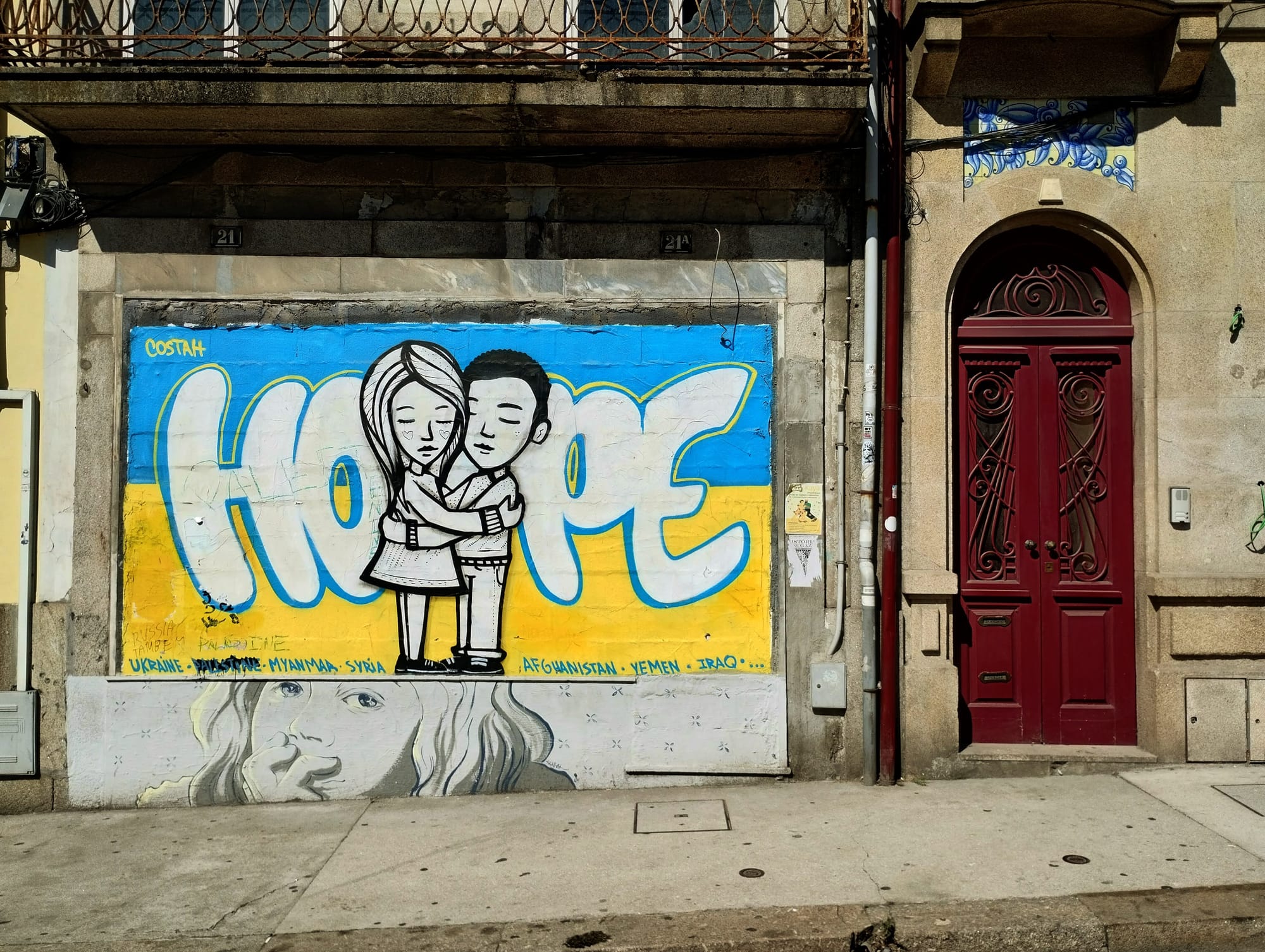 A red wooden door with decorative metal elements to the right of a mural of the Ukrainian flag featuring a man and woman hugging in front of the word "HOPE".