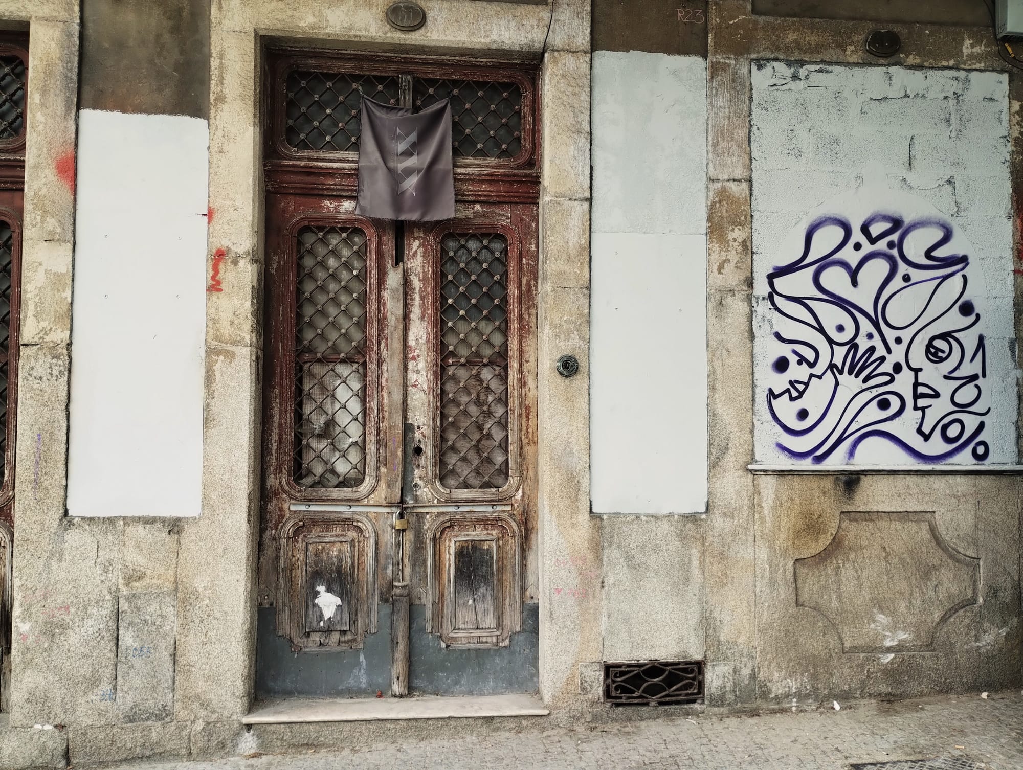 Weathered wooden double doors with wire mesh windows, a hanging banner, and graffiti on a textured stone wall facade with white painted panels.