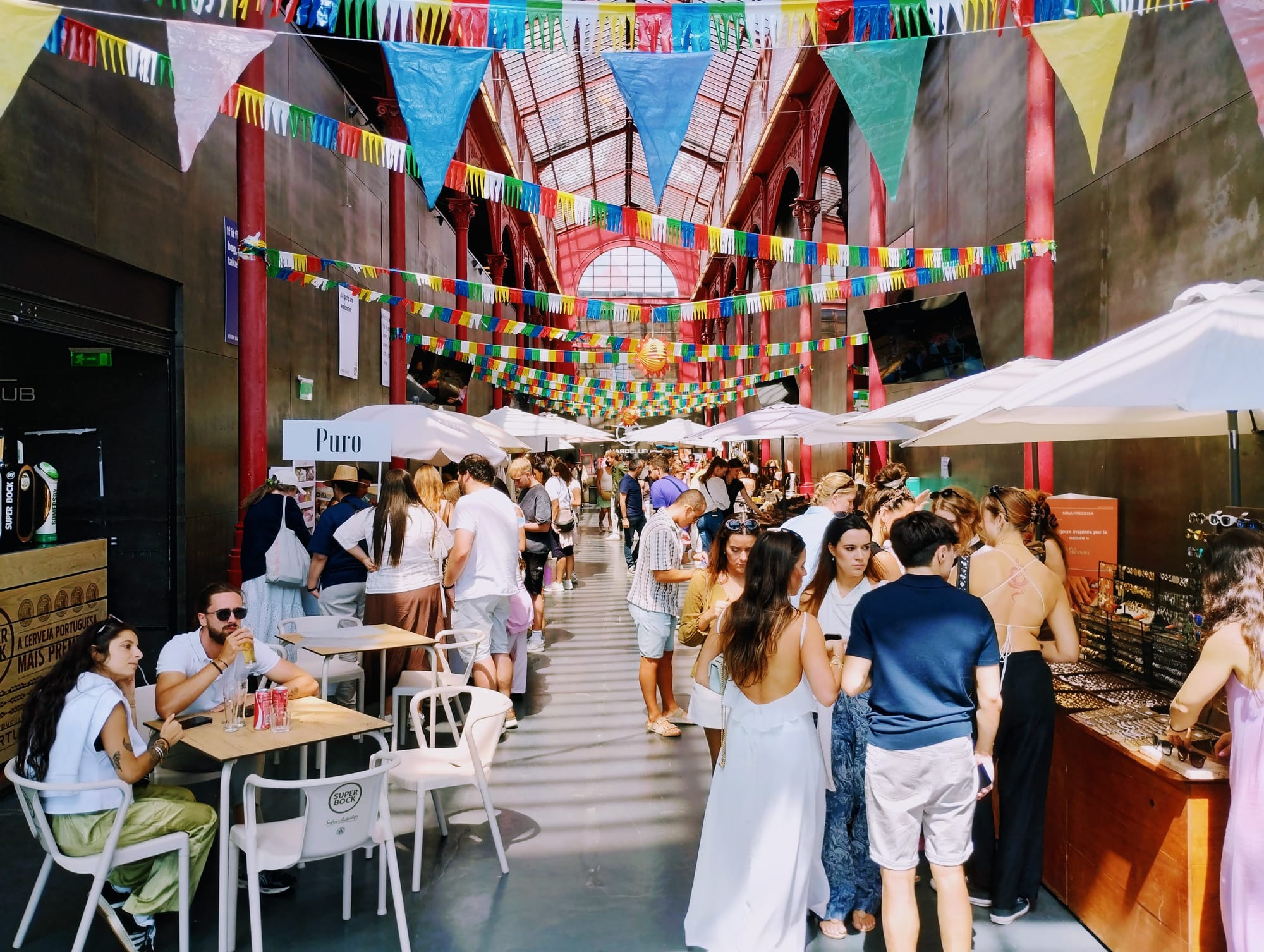 People browse market stalls and enjoy drinks under colourful bunting in a bustling, ornate, arched building.