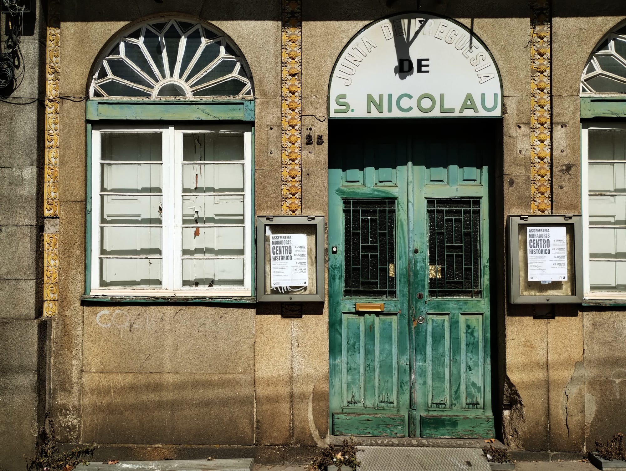 Exterior of a building featuring a pair of weathered teal double doors with wrought iron grates, flanked by a white-framed window with a sunburst design at the top and a second window. The building's stone facade includes decorative vertical tile strips and small posters beneath the window on either side of the doors. A sign above the doors reads "JUNTA DE FREGUESIA DE S. NICOLAU."