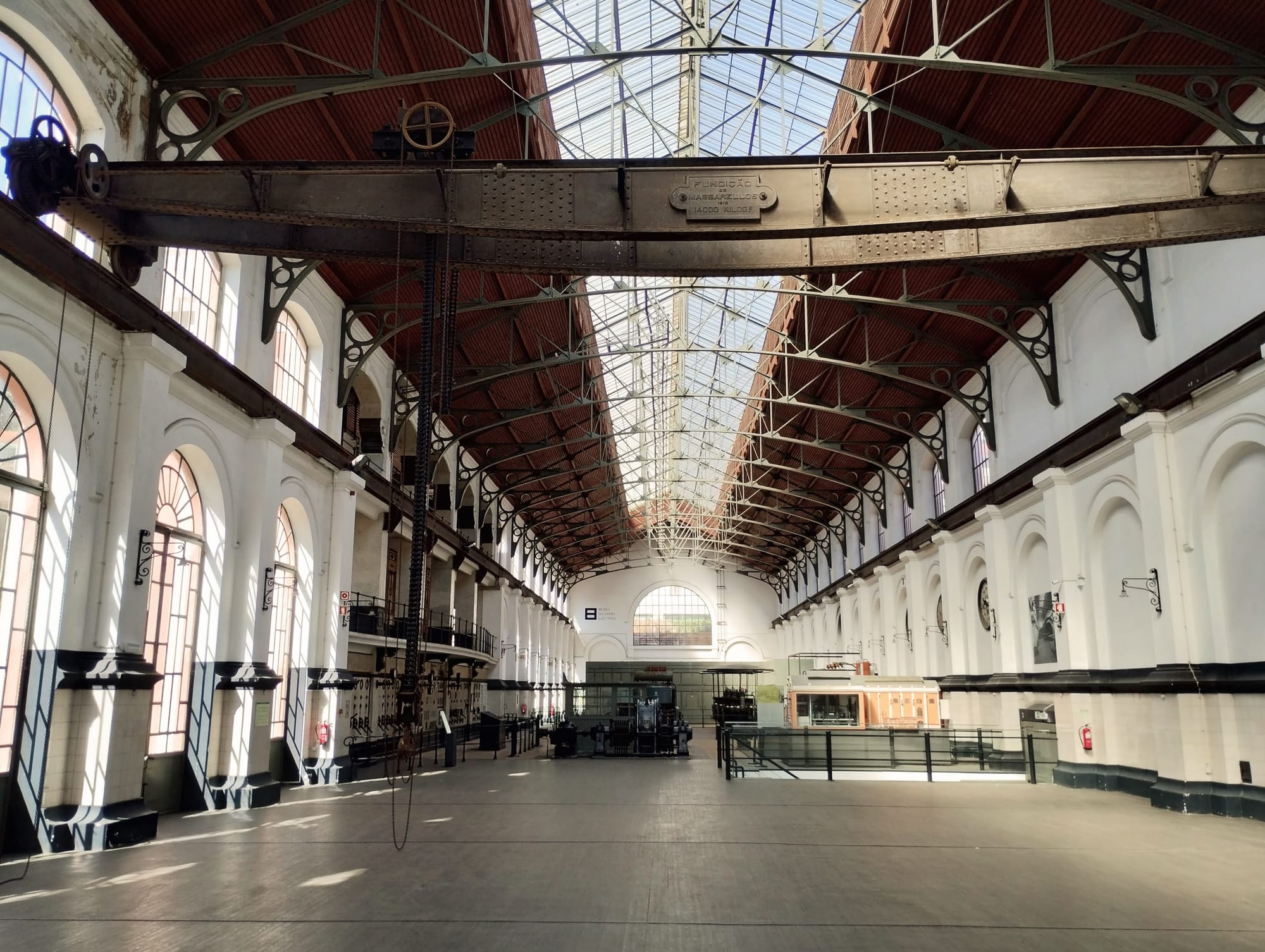 Interior view of a large industrial building with a high arched ceiling with glass skylight, exposed steel beams, and red-painted panelling. Sunlight streams through large arched windows lining the white walls on either side of a wide, empty floor.  A large metal crane hangs from the ceiling, and the background shows the building's interior machinery and installations.