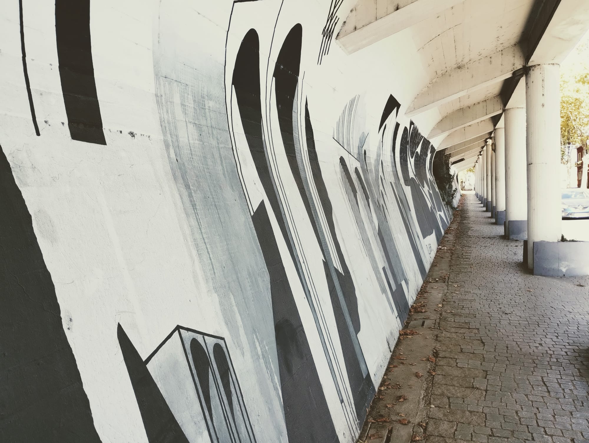 A perspective view of an underpass with a long, textured white wall featuring bold black and grey abstract artwork. The underpass has a series of white concrete pillars on the right and cobbled walkway.