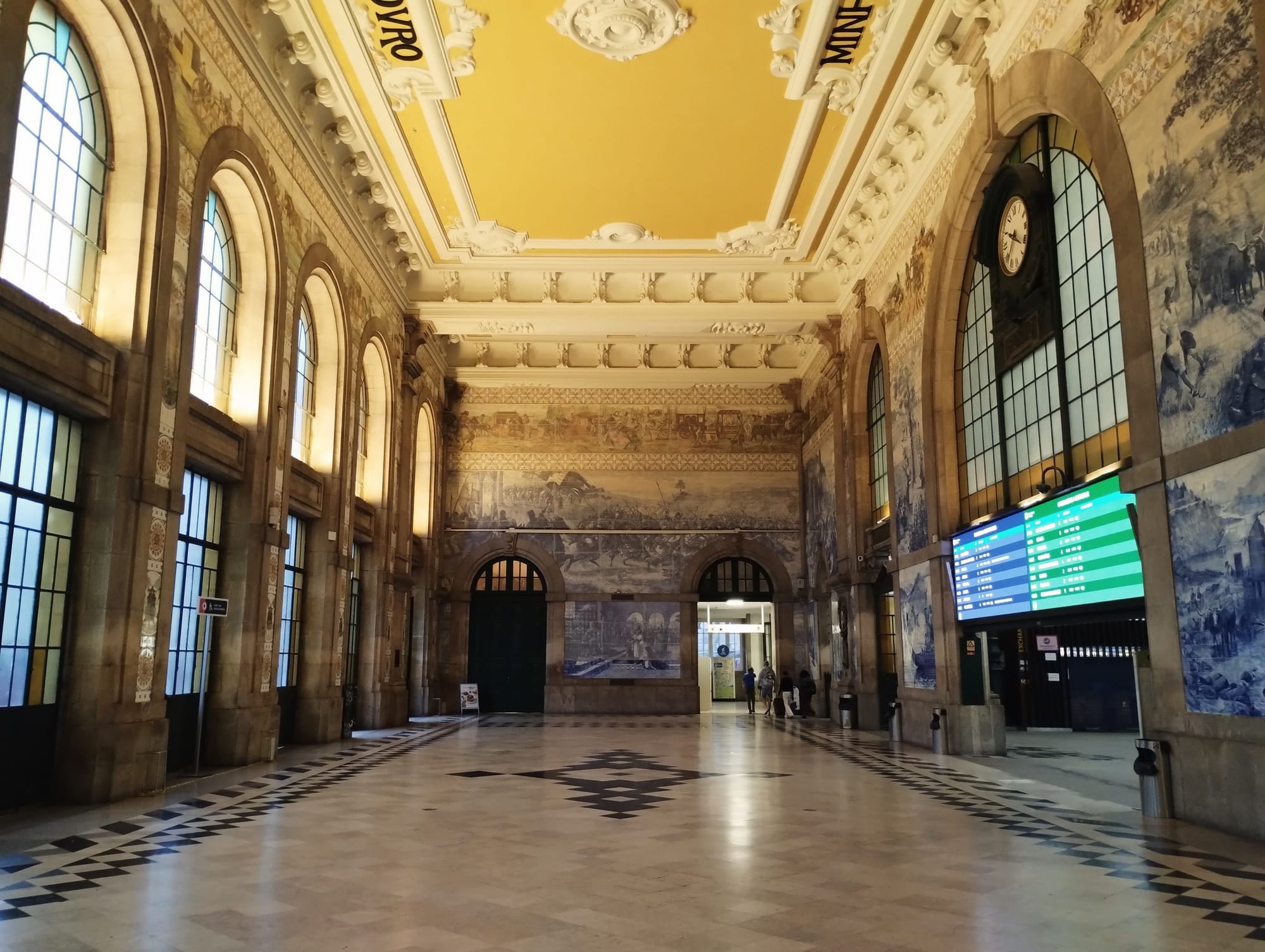 Interior of Porto Sāo Bento, featuring high arched windows, intricate ceiling moulding, and painted murals on the walls. A tiled floor in a geometric pattern.  A digital display board is visible.