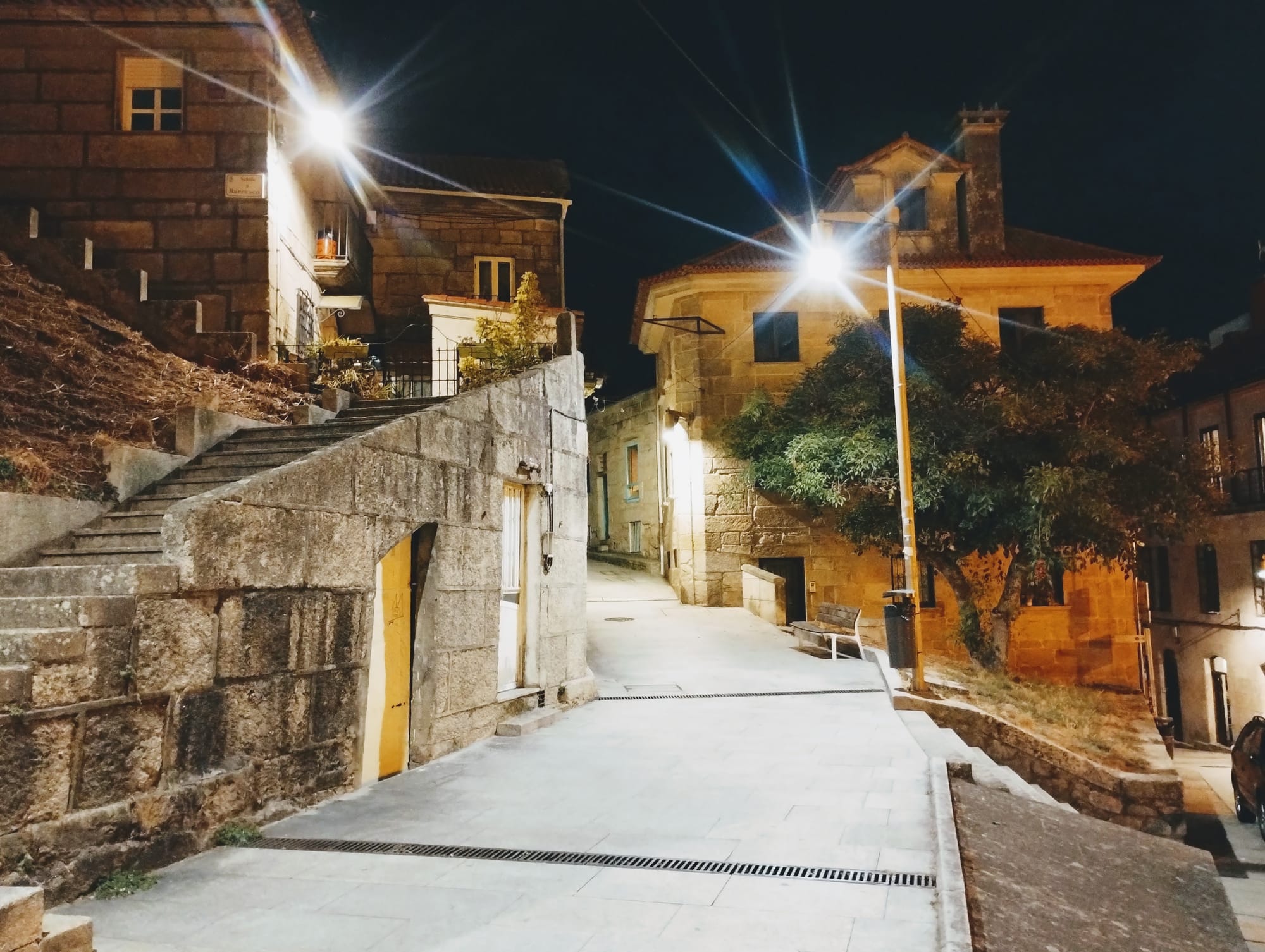 Nighttime street scene in a Vigo with stone buildings, lit by streetlights causing bright starburst effects, a staircase leading up to a building and a paved walkway.