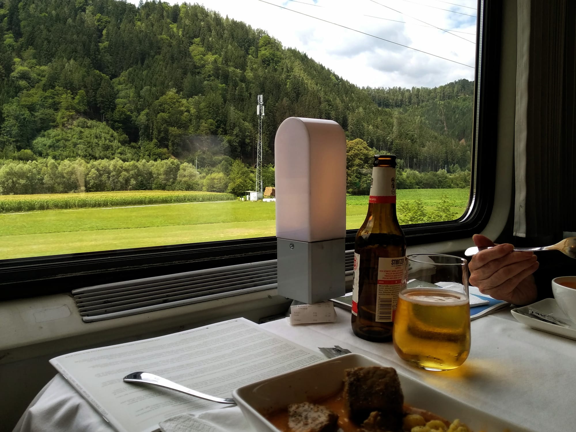 Inside a train car, a table set for a meal with a beer bottle and a glass of beer, a plate of food with meat and spaetzle, and a large window revealing a green landscape with a forest and a rudely-placed mobile phone tower.