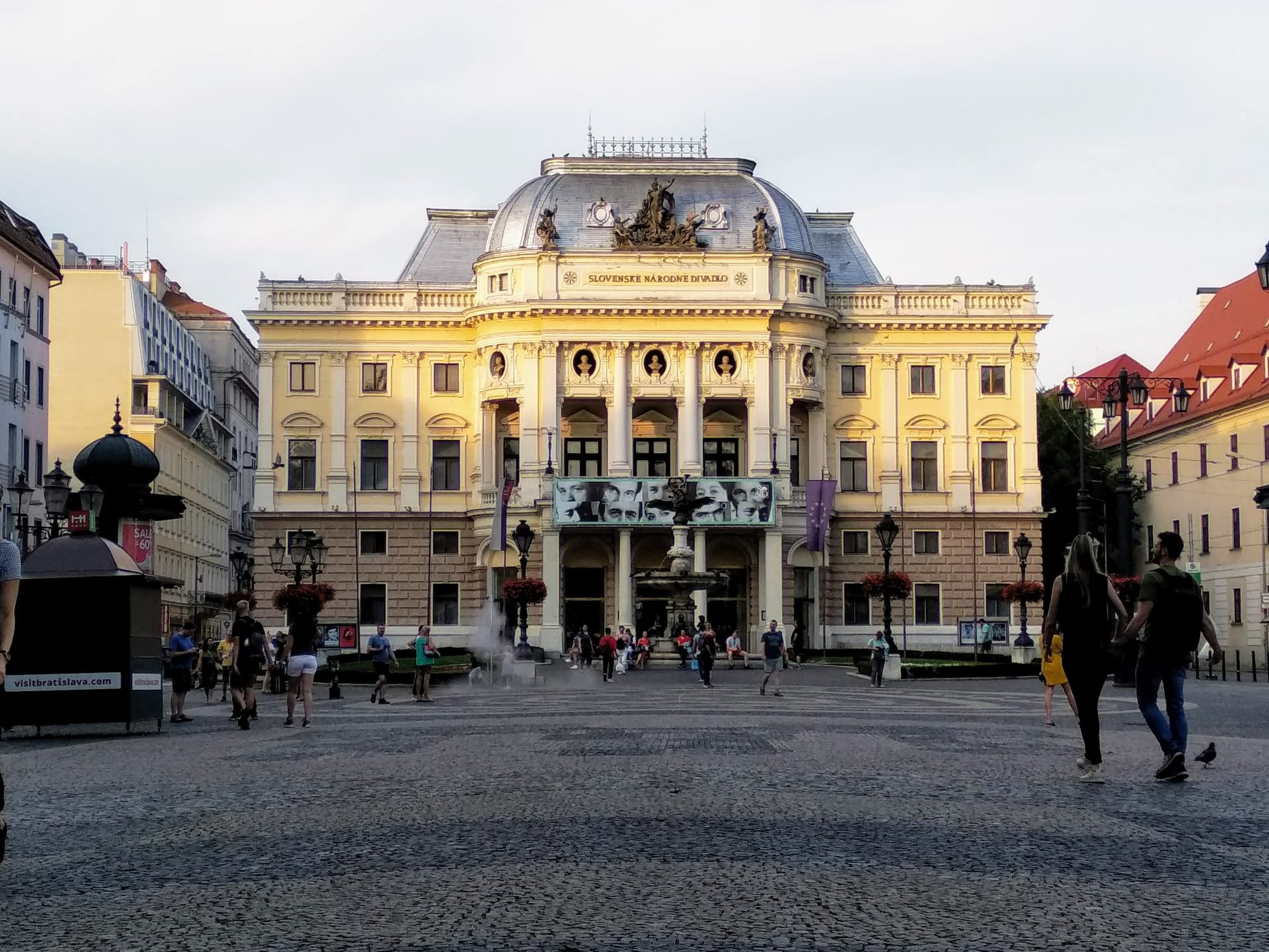 The Slovak National Theatre, a large, ornate, yellow building with a domed roof and columns, stands at the end of a cobblestone square. People are walking and standing in the plaza, and a small fountain sprays water. Buildings line the sides of the square, and the sky is overcast.
