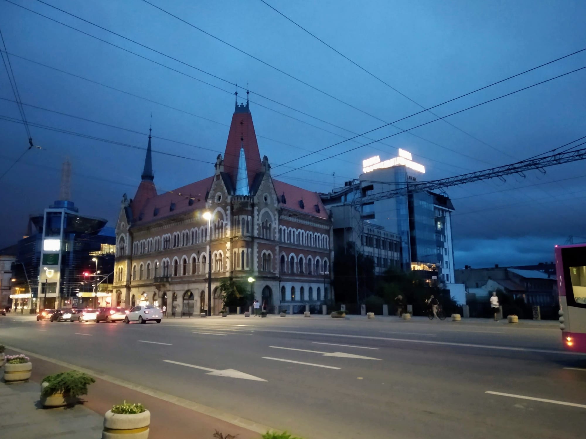 Nighttime street scene featuring a large, ornate building with a red roof, flanked by modern buildings; cars are visible on the street under a dark blue sky.