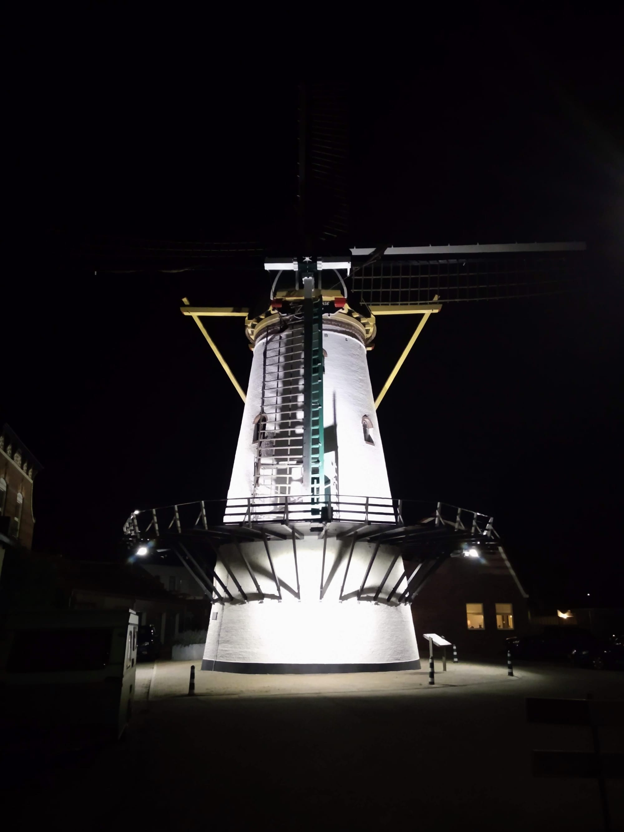 A tall white windmill illuminated at night, with blades outstretched, a ladder running up the side, and a platform at the base of the body, set against a dark sky.