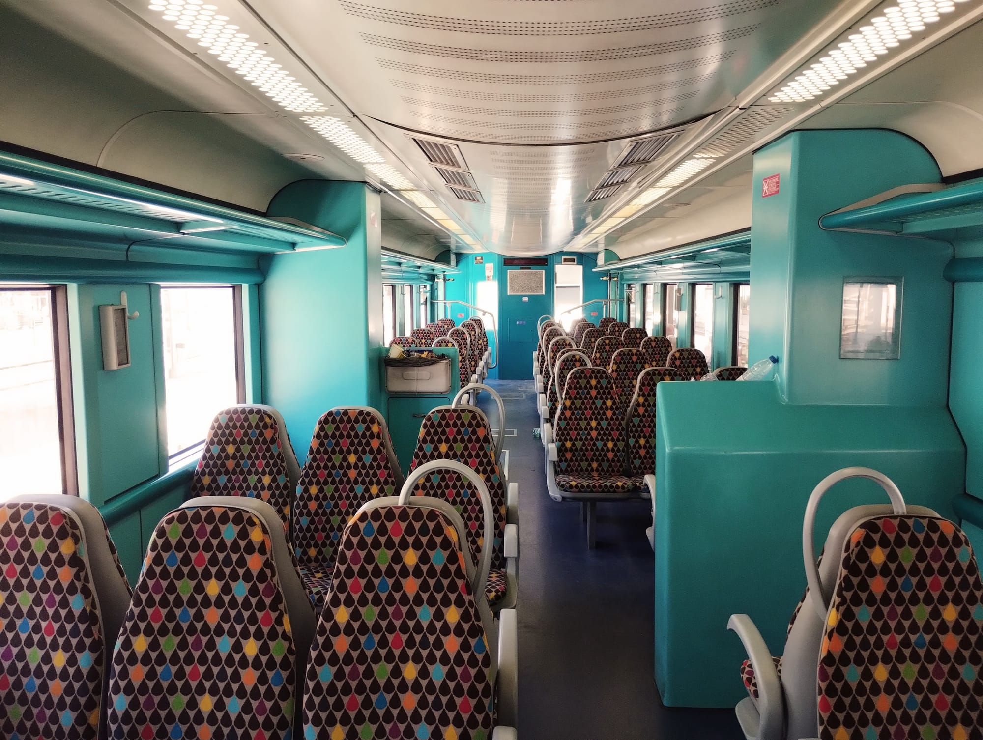 Interior of an empty passenger train car with teal walls and ceiling, and basic seats in 3+2 layout with red, blue, yellow, and black tear motif on beige moquette.