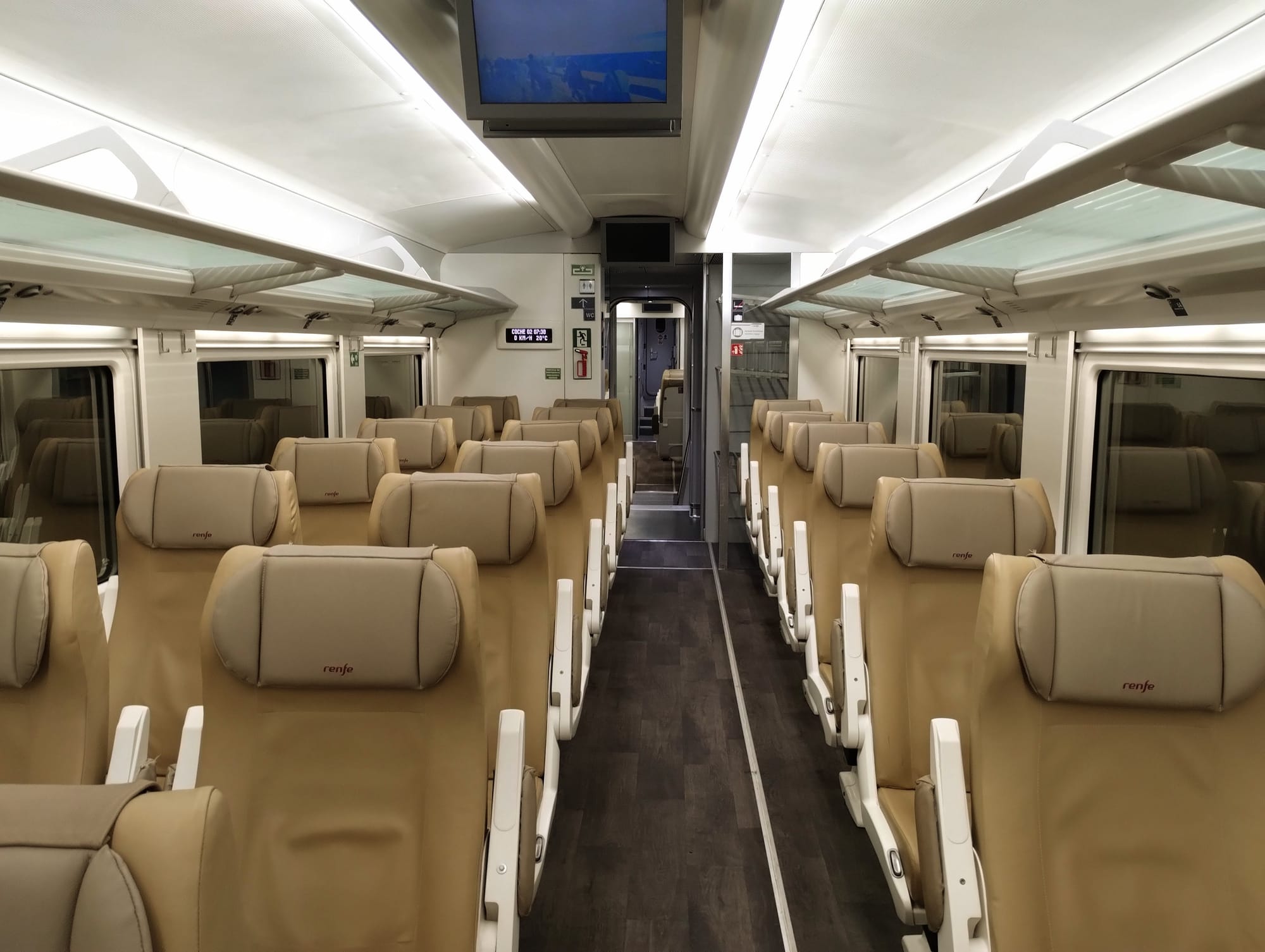 Interior view of an empty passenger train featuring rows of tan leather seats with headrests branded with "renfe".  The car has overhead luggage racks, windows, and a central aisle with a door at the end.