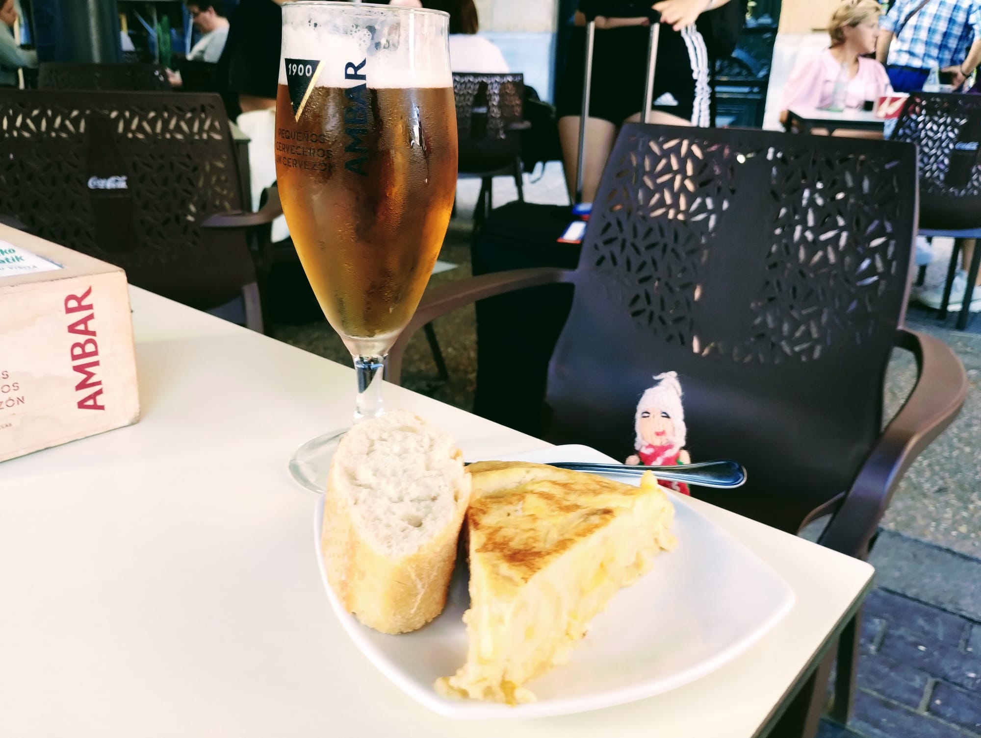 An inebriated finger puppet contemplates a glass of Ambar beer at a white table set with a slice of Spanish tortilla, and a piece of baguette.
