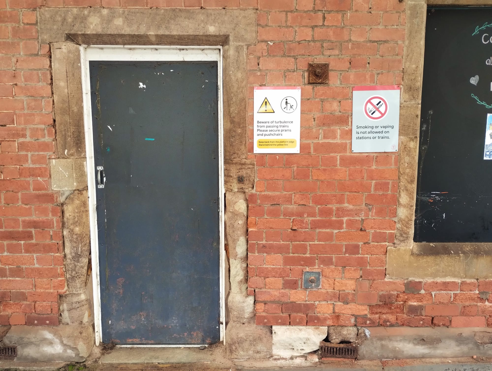 An brick wall with a dark blue metal door. The door is centered, slightly off-white, and has a handle. To the right of the door are two signs: the first reads "Beware of turbulence from passing trains. Please secure prams and pushchairs. Keep back from the platform edge. Stand behind the yellow line" with a warning sign and an image of a person with a stroller. The second sign to the right states "Smoking or vaping is not allowed on stations or trains."
