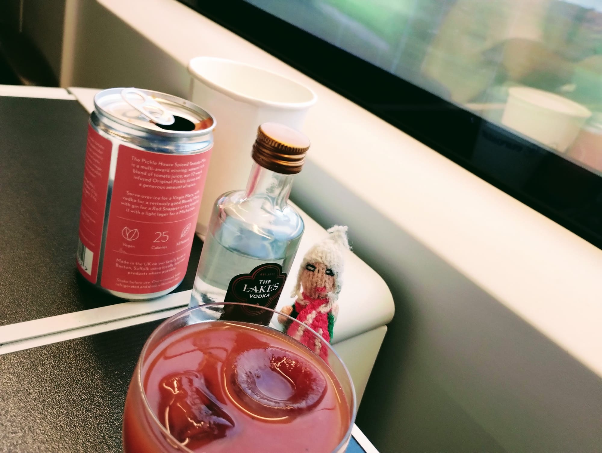 Bloody Mary cocktail components on a train seat table. The ingredients are a can of 'The Pickle House Spiced Tomato Mix' and a miniature bottle of 'The Lakes Vodka'. A knitted finger puppet lush surveys the glass containing a Bloody Mary with two ice cubes.