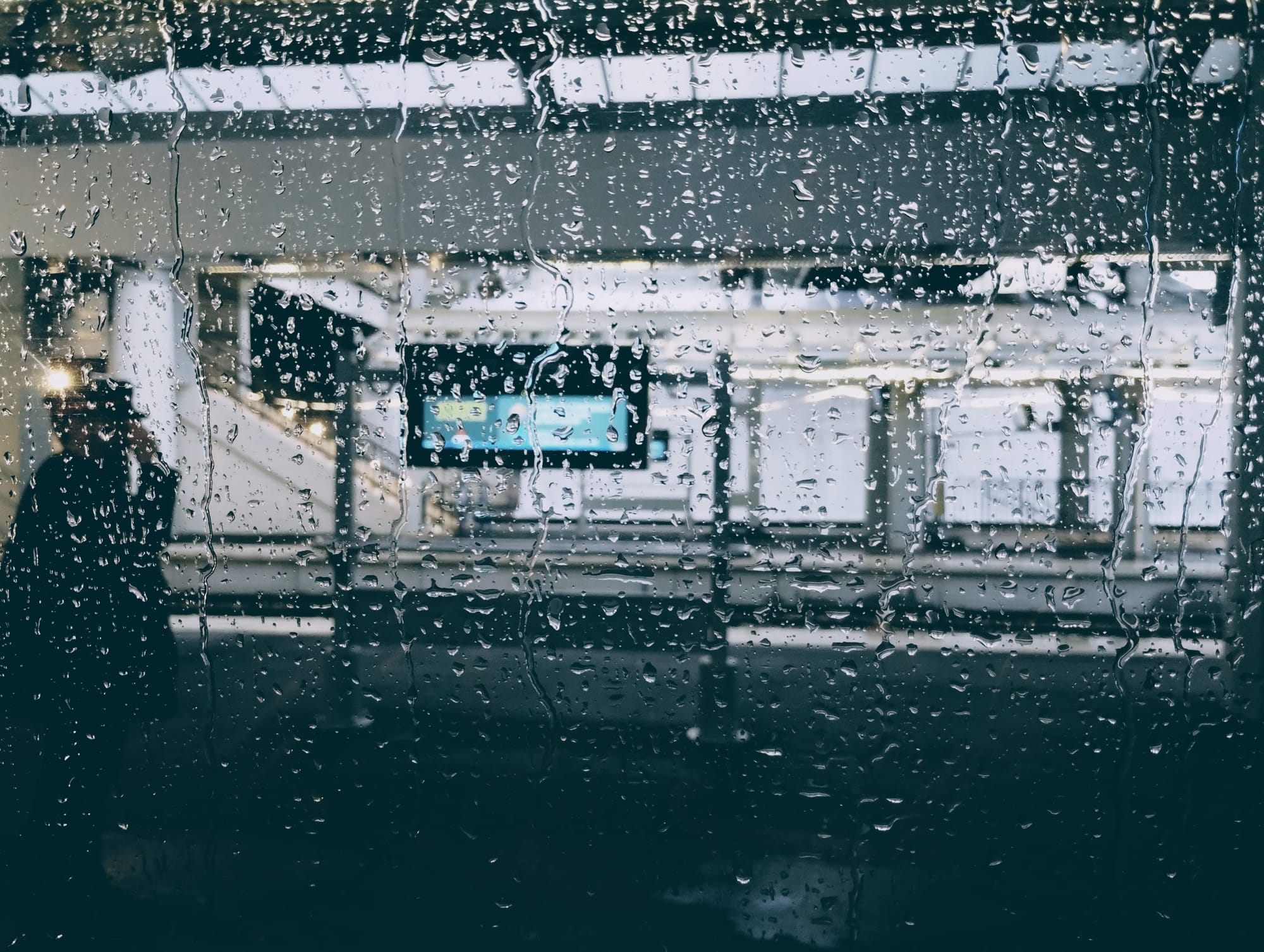 View through a rain-streaked window of a platform in Limoges Benedictins. Raindrops obscure the scene, partially obscuring a person in silhouette on the left and a digital display screen in the centre.