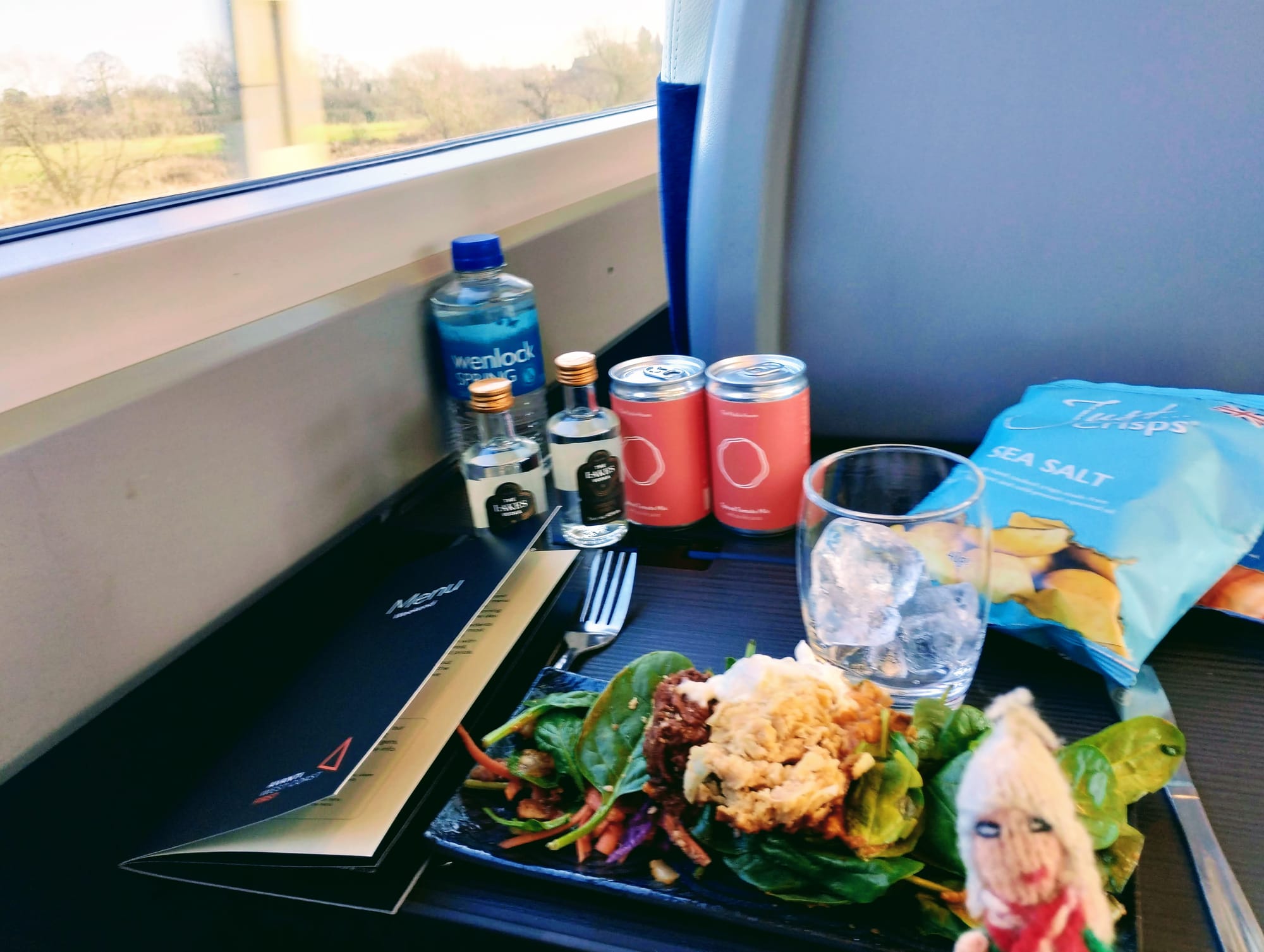 A small Peruvian finger puppet in front of a tray on a train table containing meal of salad, a glass with ice, crisps, two bottles of vodka and two cans of tomato juice. A window with a view of a green landscape is visible.