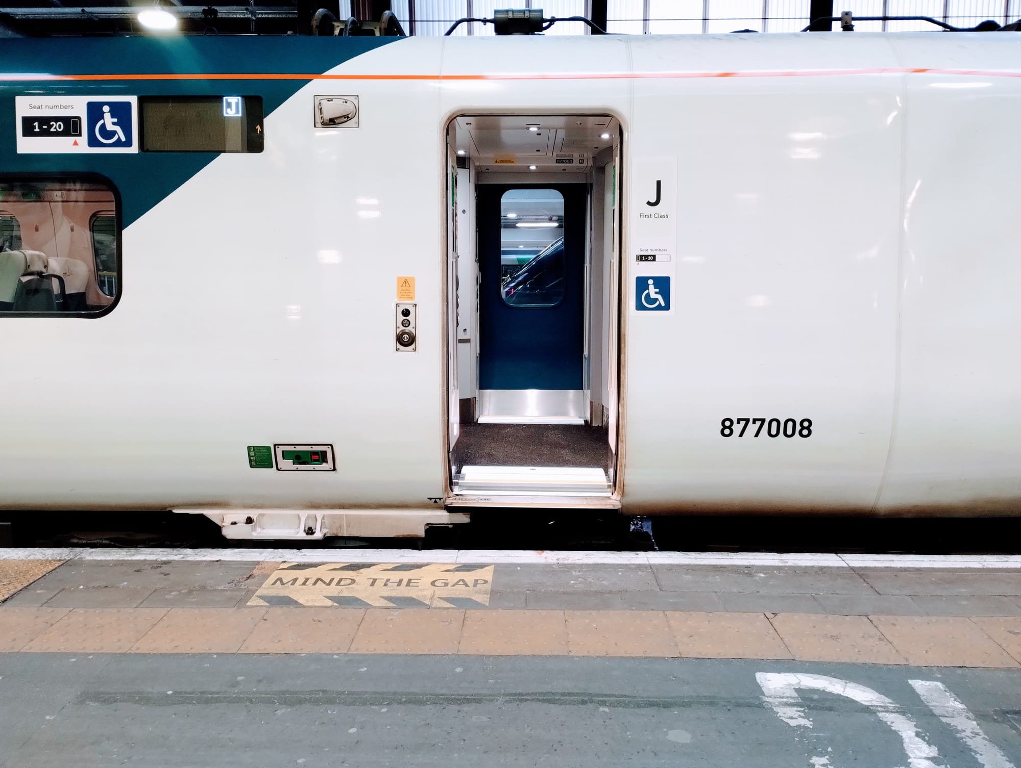 Open train door revealing interior, next to signs for first class and accessible seating, on a station platform with "Mind the Gap" warning. The pointy end of another train can be seen through the window on the other side.