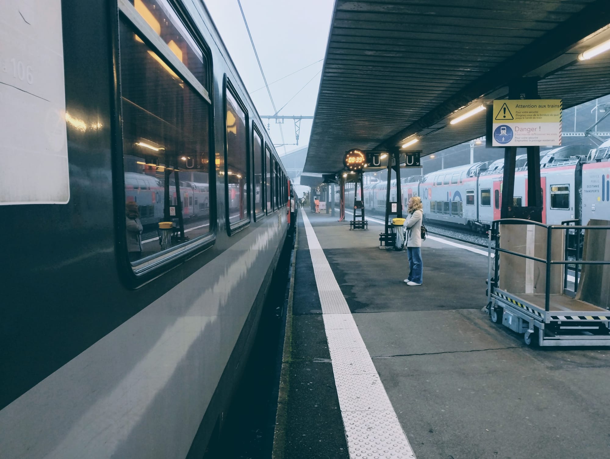 Train at a station platform on a foggy morning, woman in casual clothes stands near train tracks, with overhead canopy, signage, and a train whose carriages disappear to the horizon.