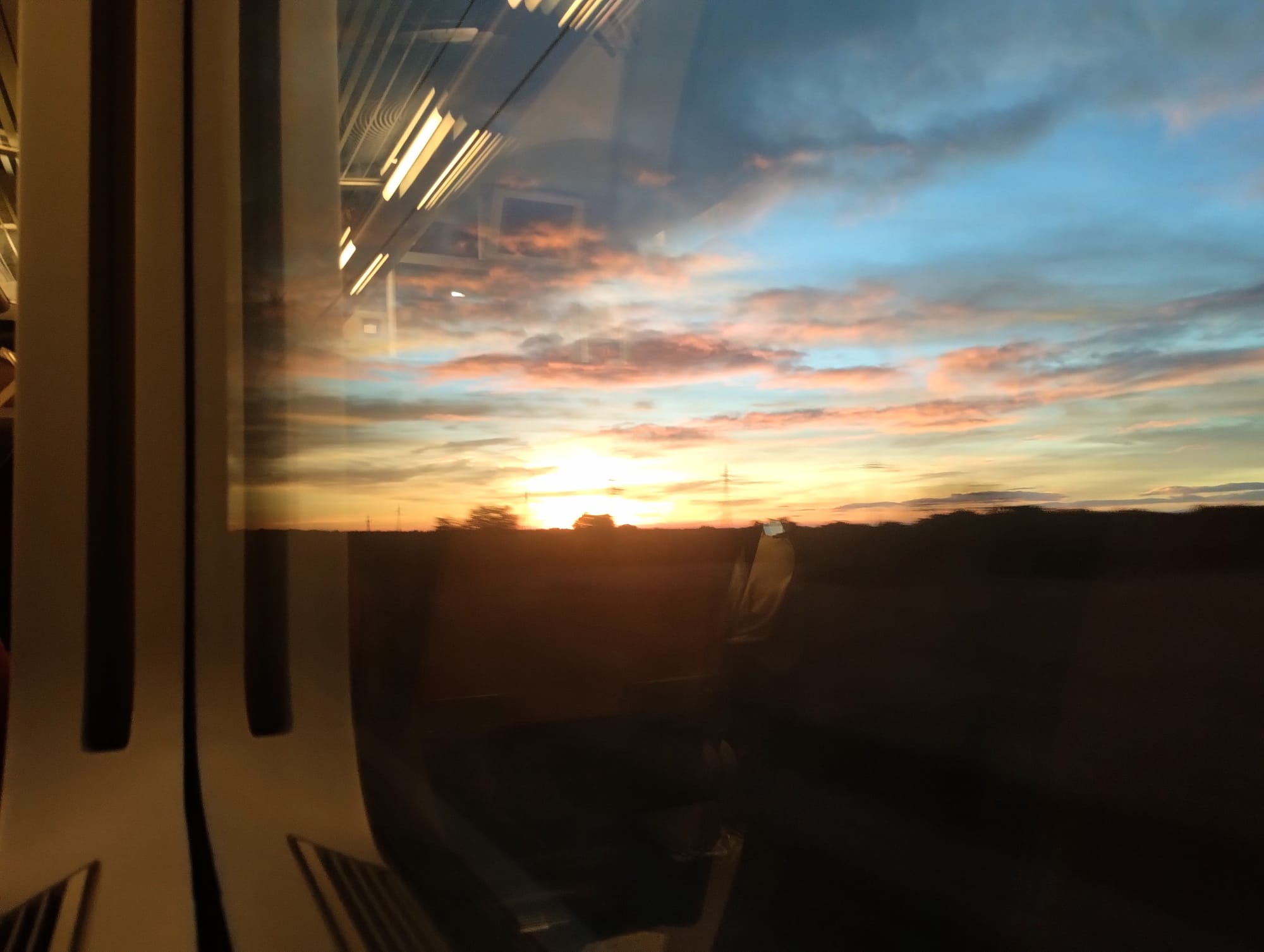 View through a train window of a sunset with an orange and blue sky. Reflected in the glass, with the interior of the train in the foreground with vertical panels.