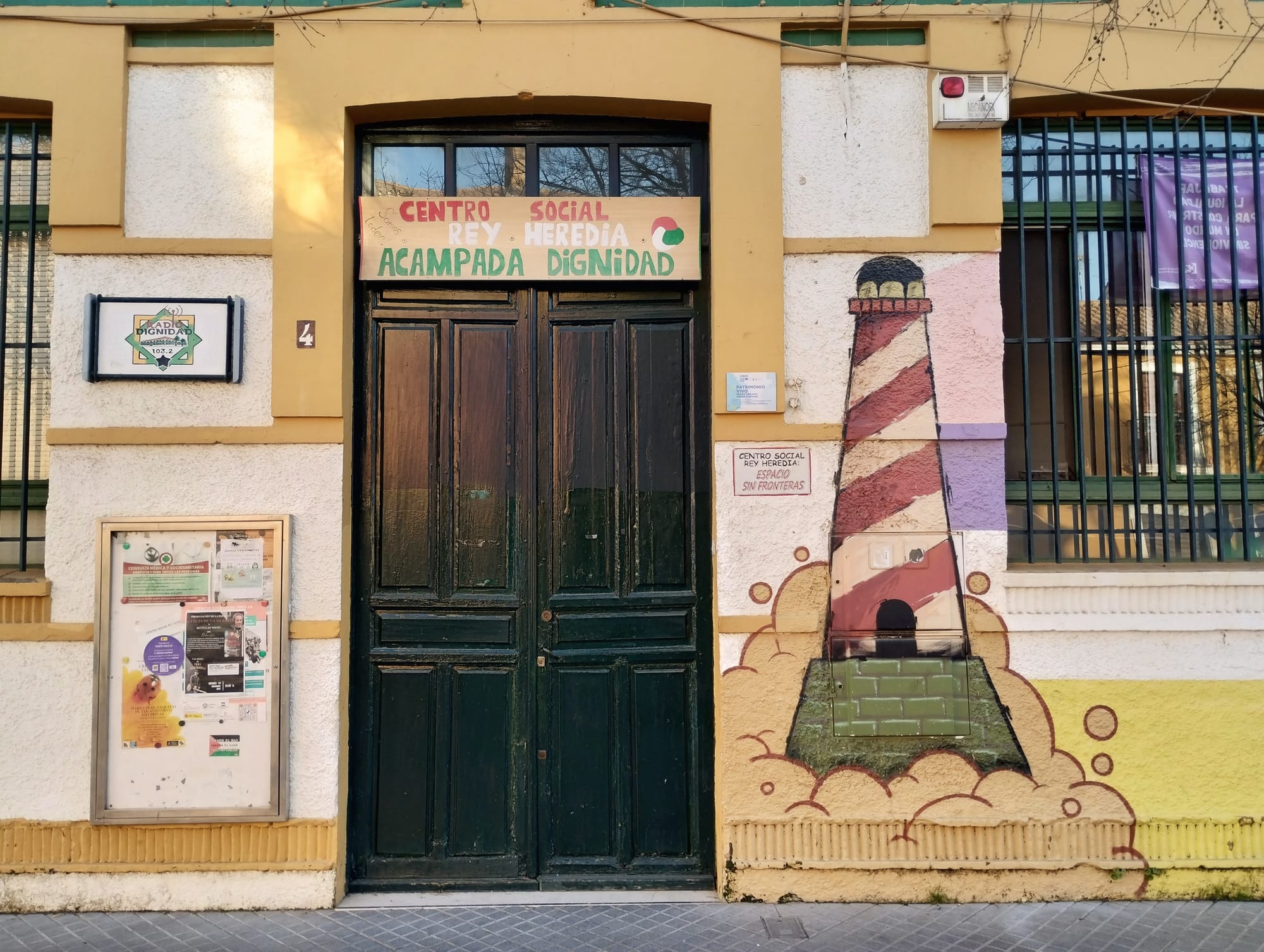 Façade of a building with a dark green double door and a painted red and white striped lighthouse mural. A banner above the door reads "CENTRO SOCIAL REY HEREDIA, ACAMPADA DIGNIDAD." To the left of the door is a bulletin board with flyers. Other details include windows with bars, street number 4, and an advert for Radio Dignidad.