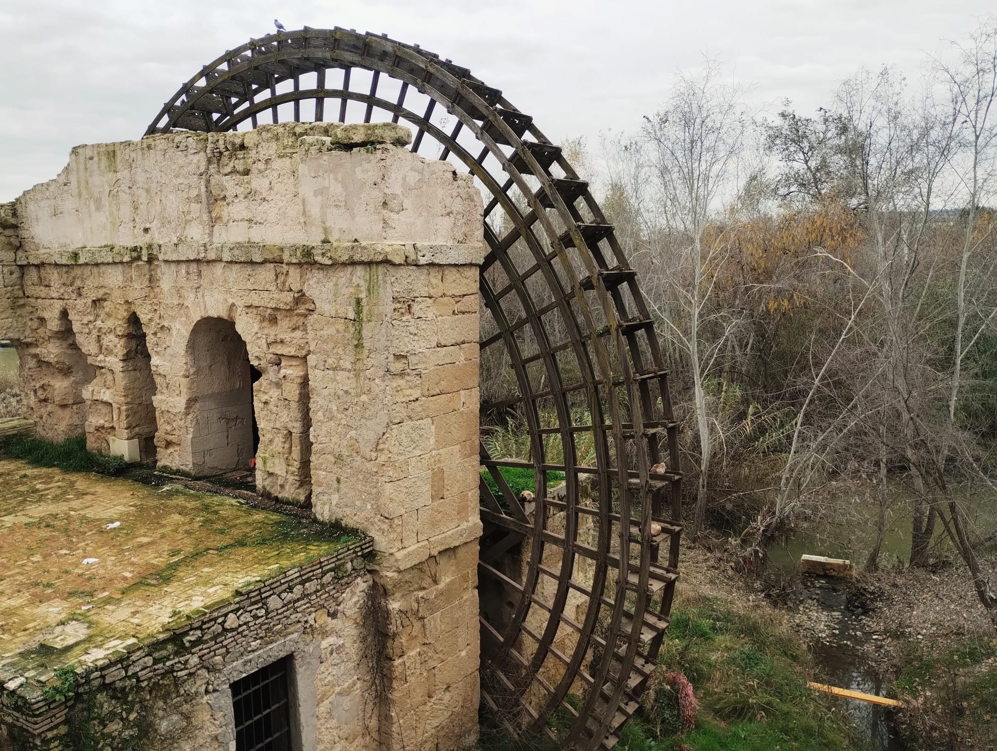 Ruined stone building with three arched openings, next to a large, weathered wooden water wheel, against a backdrop of bare trees and a cloudy sky.