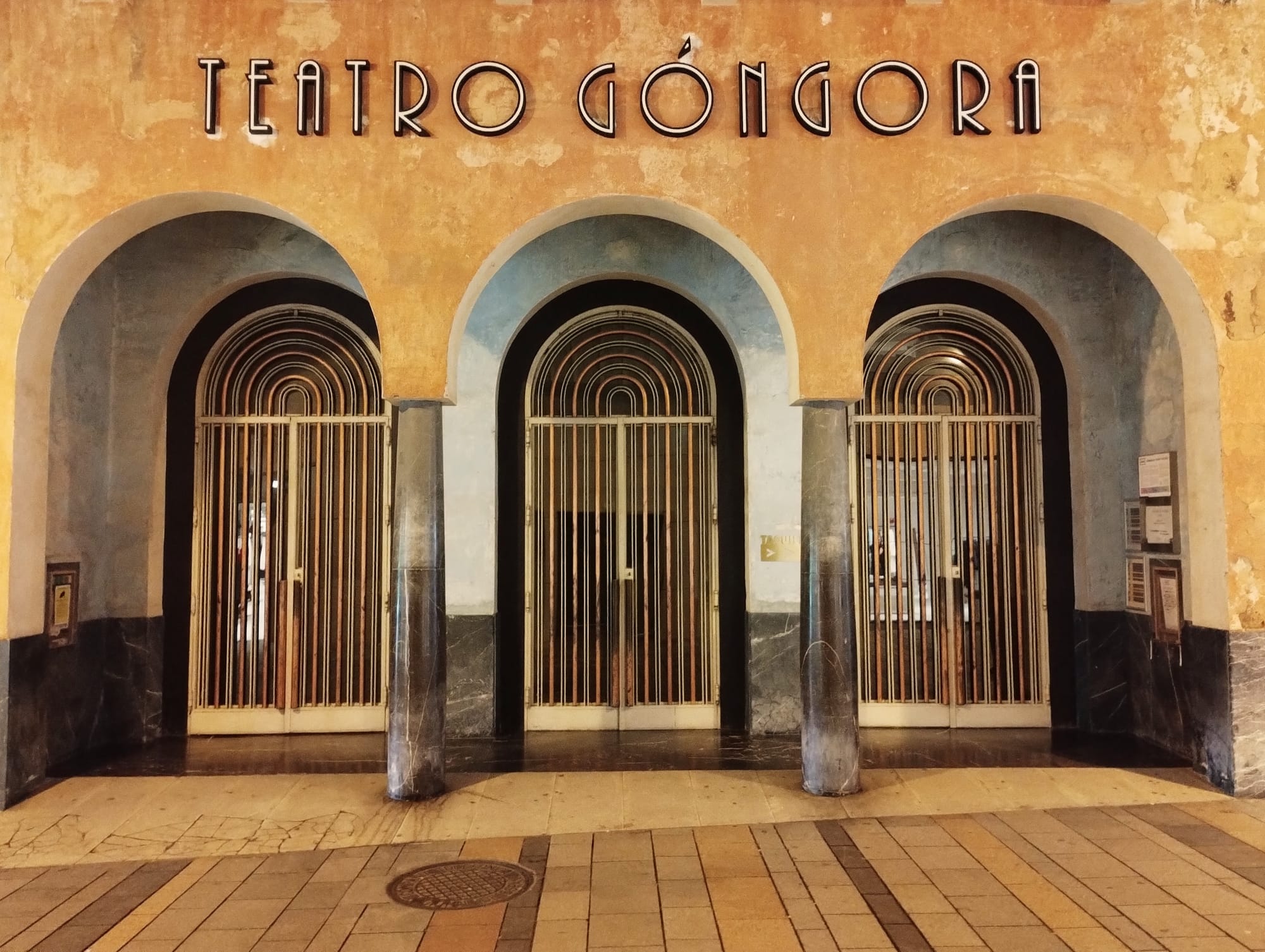 The exterior of the Teatro Góngora, featuring three arched entryways with ornate metal gate doors, centered beneath the theatre's name in bold letters against a textured, amber facade.