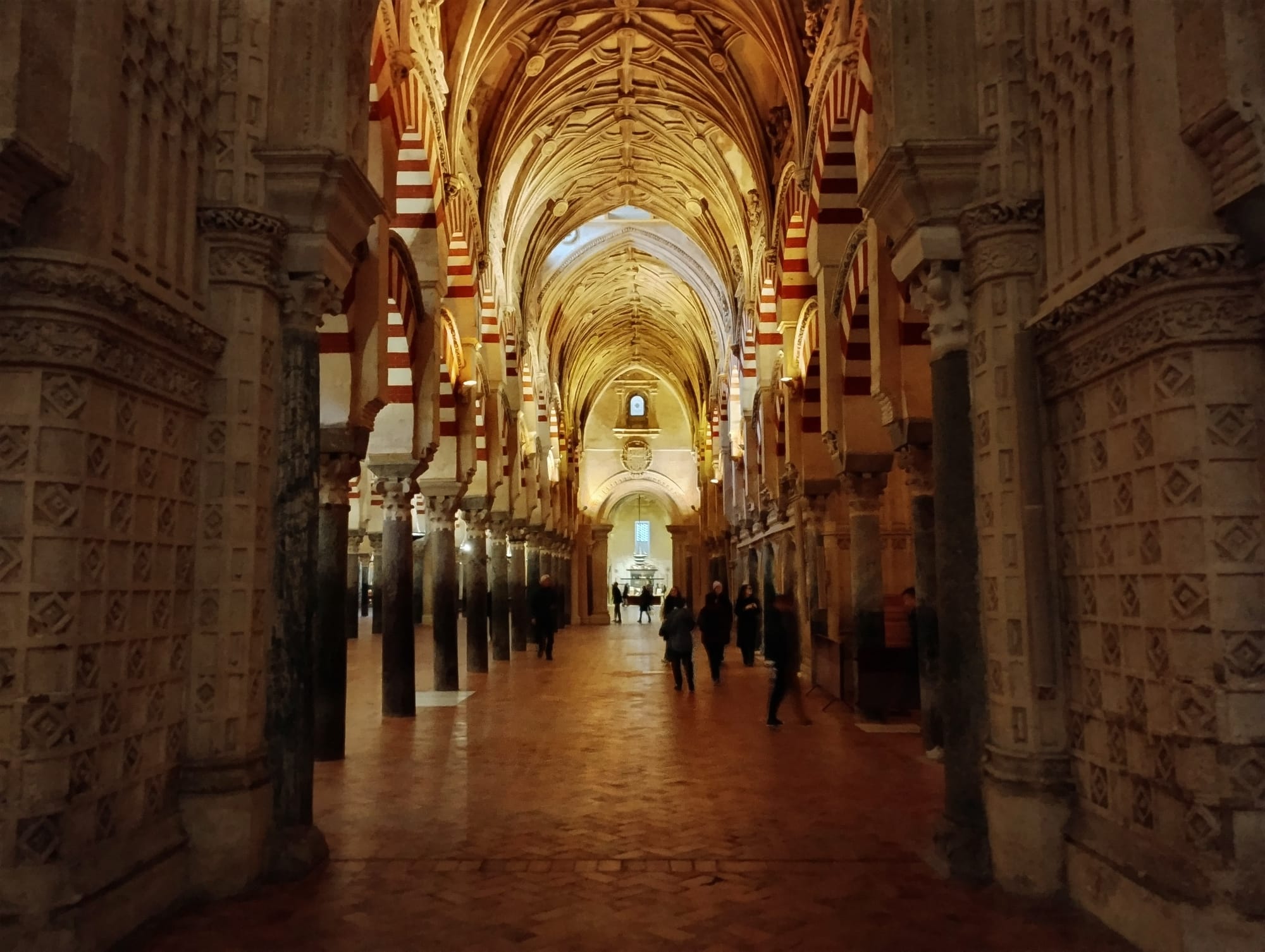 Interior view of the Mezquita-Cathedral of Cordoba, Spain, featuring a long nave with a series of archways supported by alternating red and white striped arches on marble columns, leading to a distant archway and a small group of people. The ceiling is intricately carved in a gothic style, and the floor is made of reddish-brown brick.