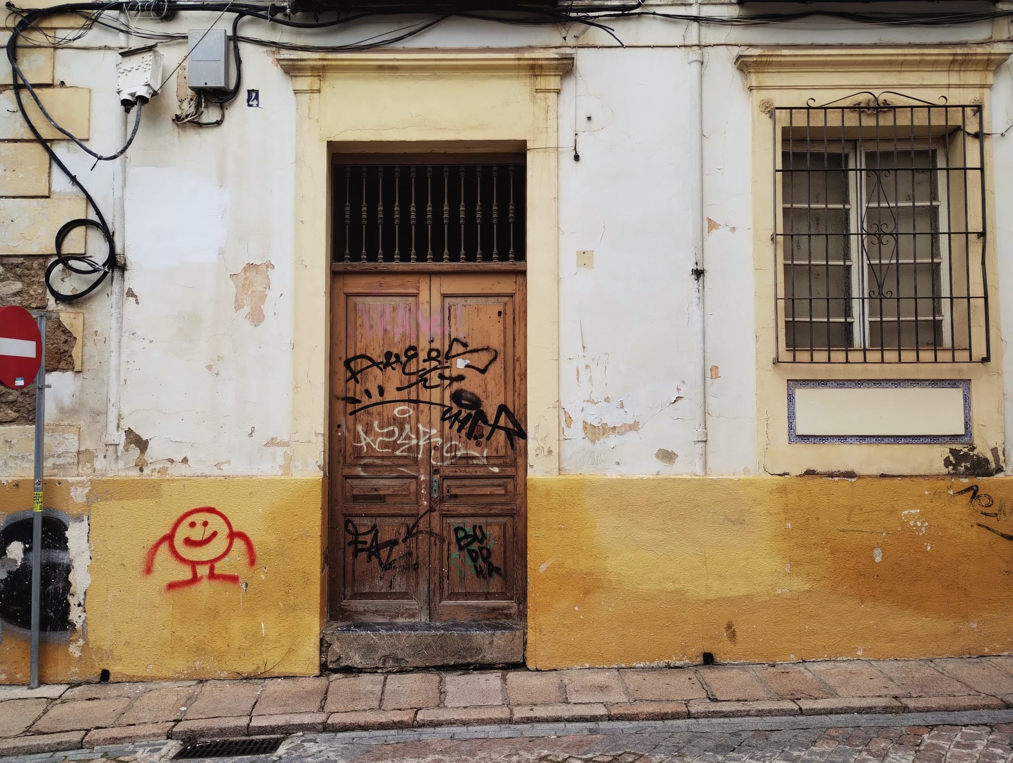 Exterior view of a weathered building facade with a wooden door covered in graffiti, a barred window, and a painted smiley face. The building is painted cream and yellow, showing signs of wear with peeling paint and exposed brick. Electrical wires and a no-entry sign are present.