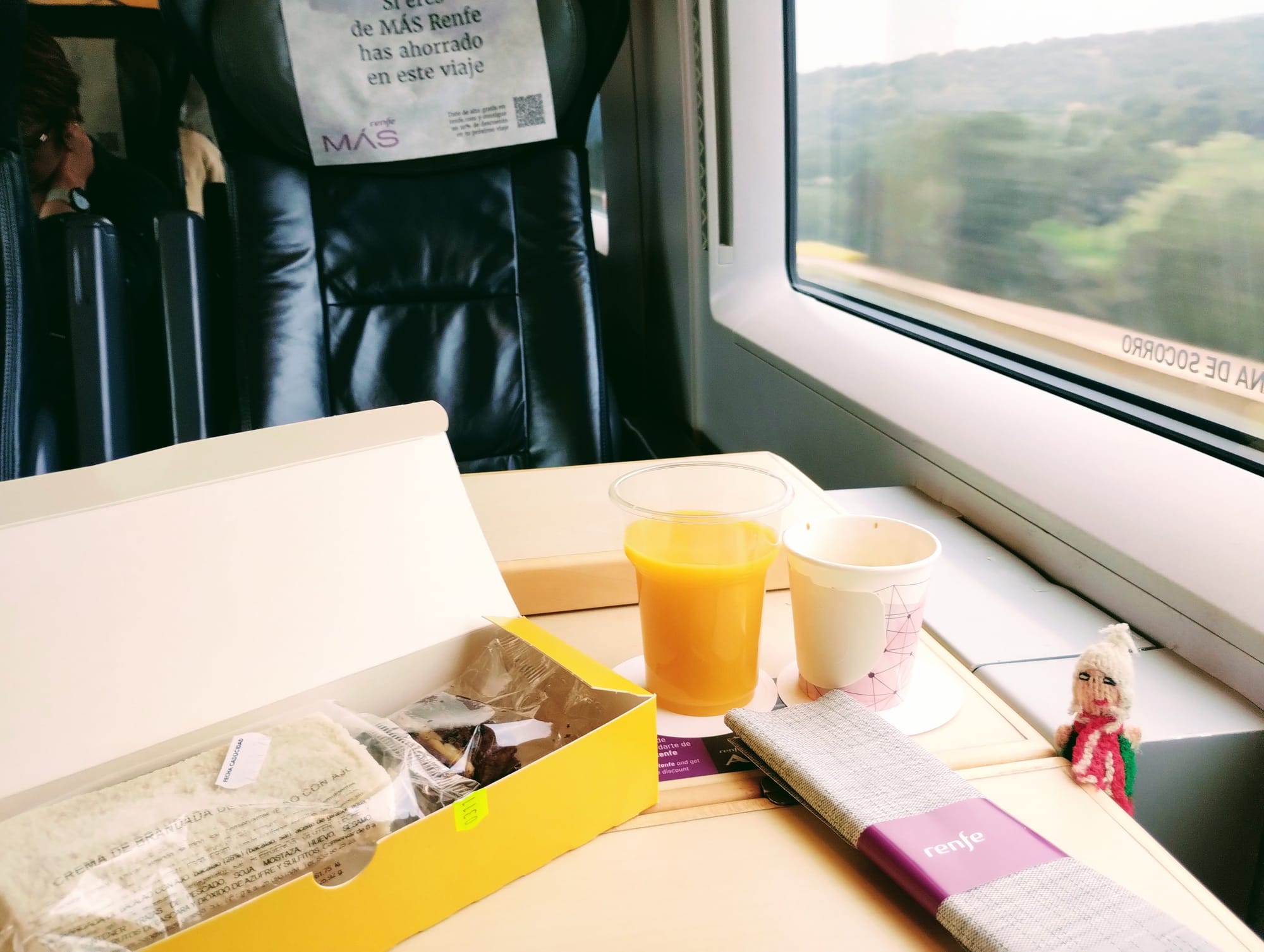 A meal on a train features a meal box, juice, coffee, and an adorable Peruvian finger puppet looking on. Outside the window, a blurred landscape speeds past.