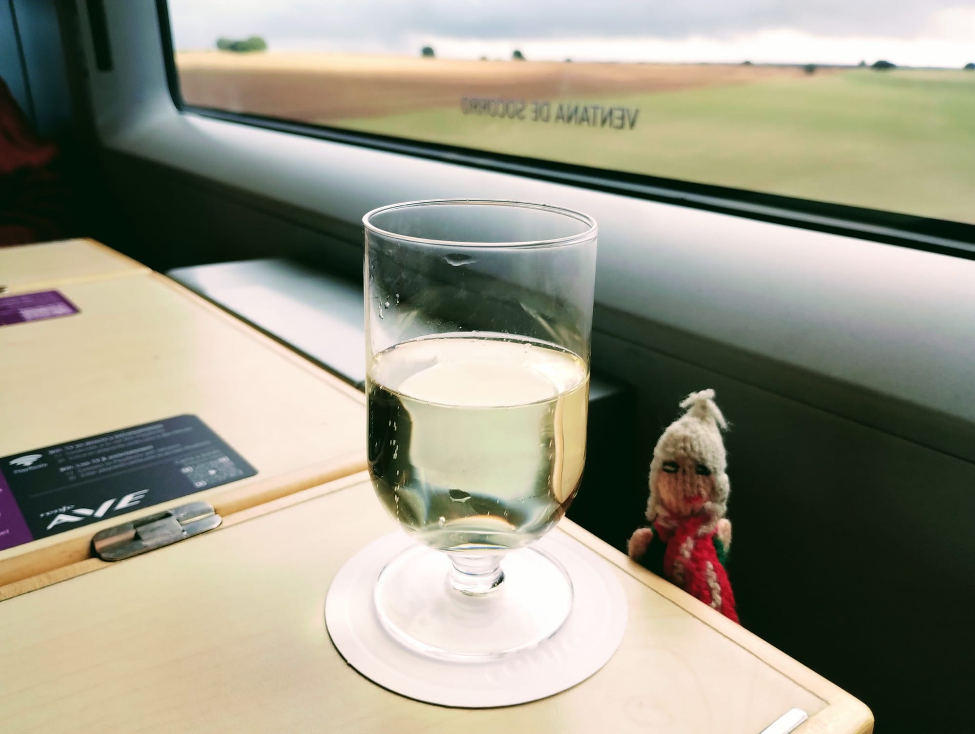 A stemmed glass filled with sparkling white wine rests on a white coaster on a light wood table, inside a train car with a view of a green field under a cloudy sky. A blurry Peruvian finger puppet waits eagerly for its first sip.