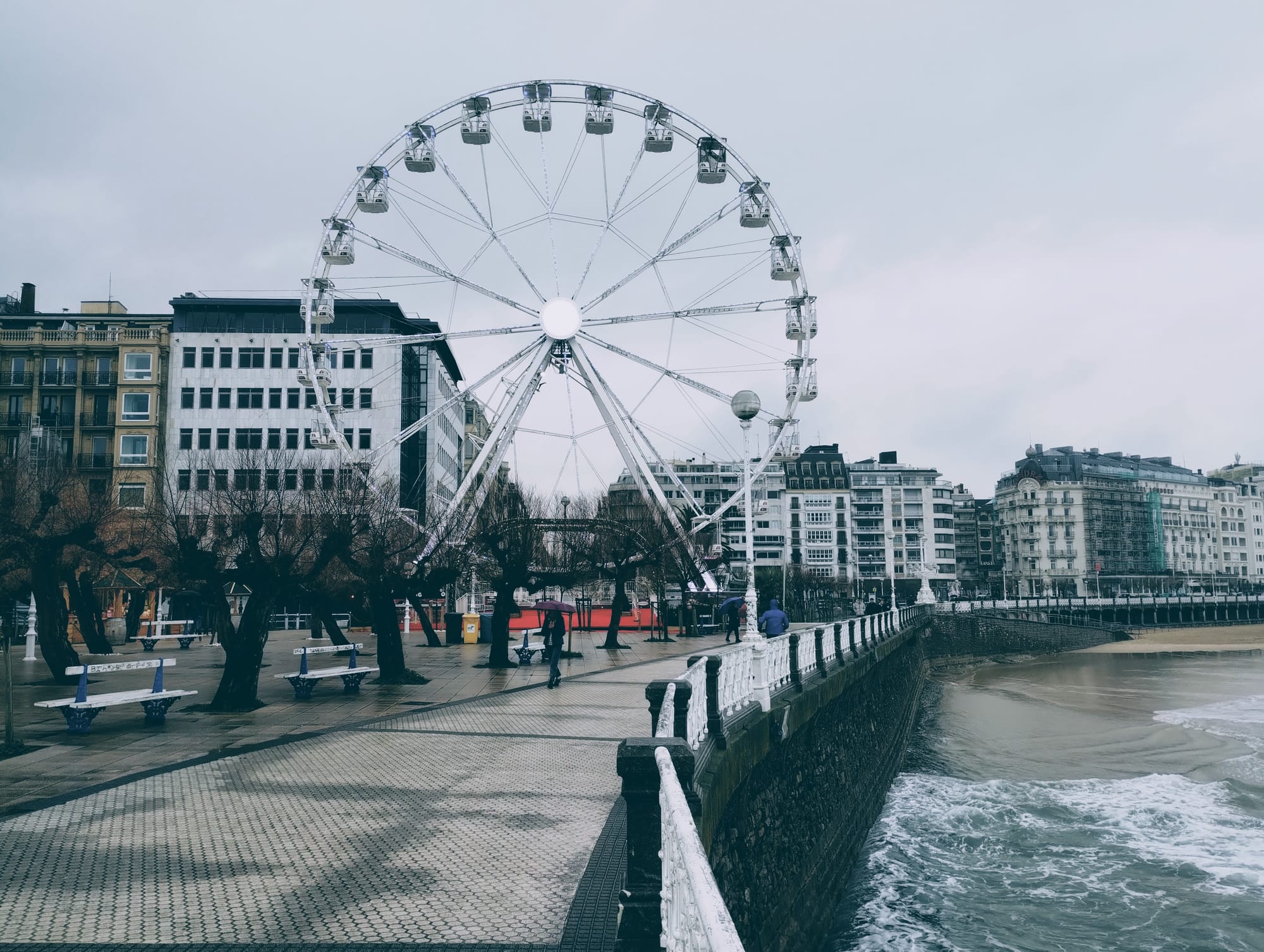 A large white Ferris wheel overlooking a beach promenade in San Sebastian on an overcast day. Buildings line the street behind the wheel, while a stone wall and white railing border the waves. Bare trees, benches, and people with umbrellas dot the foreground.