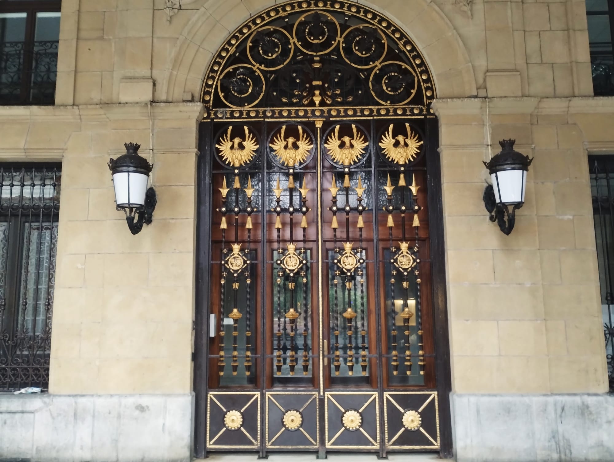 Ornate double doors with gold and black detailing and arched top, flanked by decorative wall-mounted lamps.