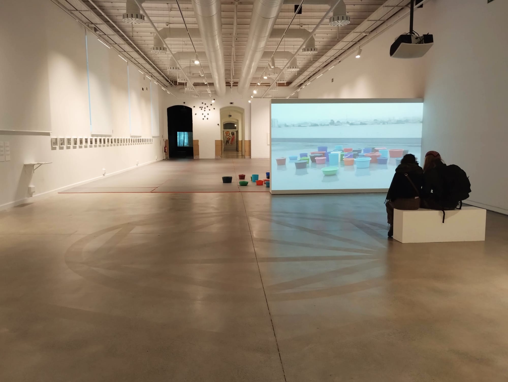 Two people seated on a white rectangular block observe a projection of buckets in the rain. Behind the screen, there are buckets on the floor. I can't make it any more interesting than it isn't.