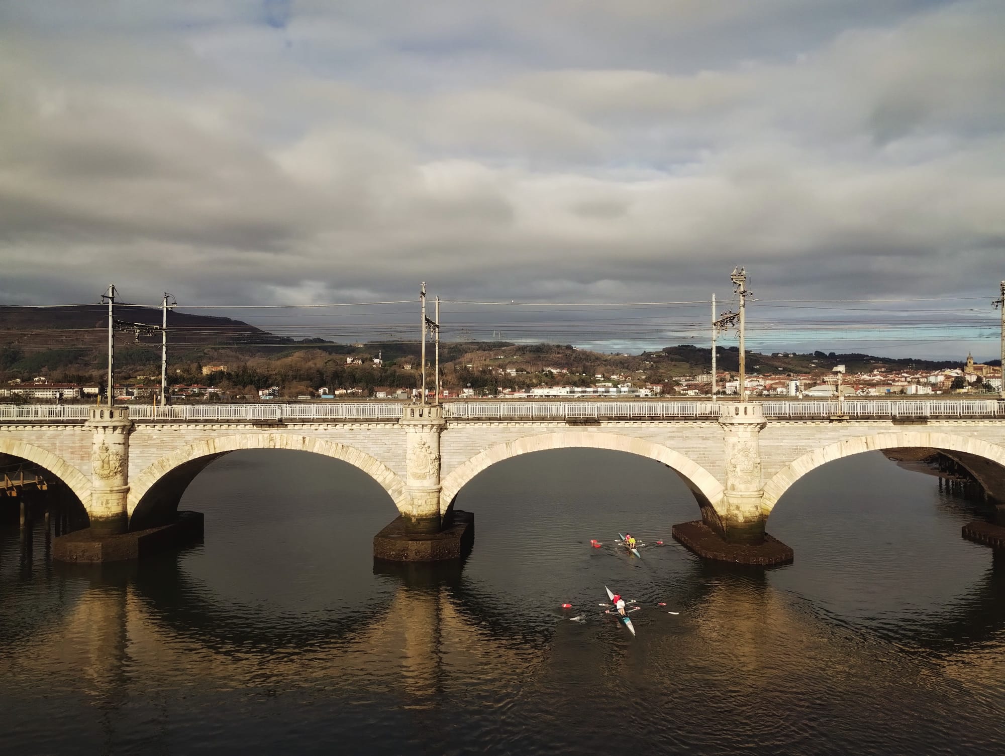 A stone arch bridge spanning a river, with two rowing teams in the water below. The bridge is composed of three large arches, with elaborate details on the piers and a railway line above. The river water reflects the bridge and cloudy sky.  The backdrop features a distant town nestled on a hillside under a cloudy sky.