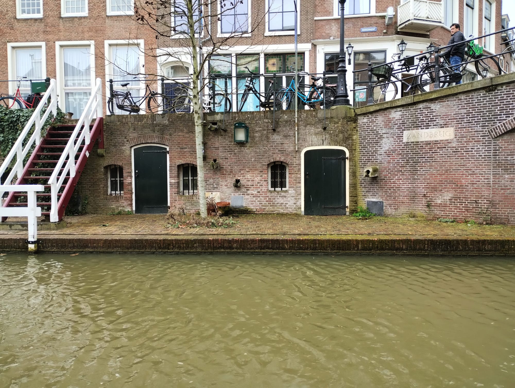 A canal-level view of a brick wall with entrances into cellars. The canal water is a murky brown. On the left, a white and red stairway with a railing leads up to street level, where bikes are leaned against railings in front of houses.