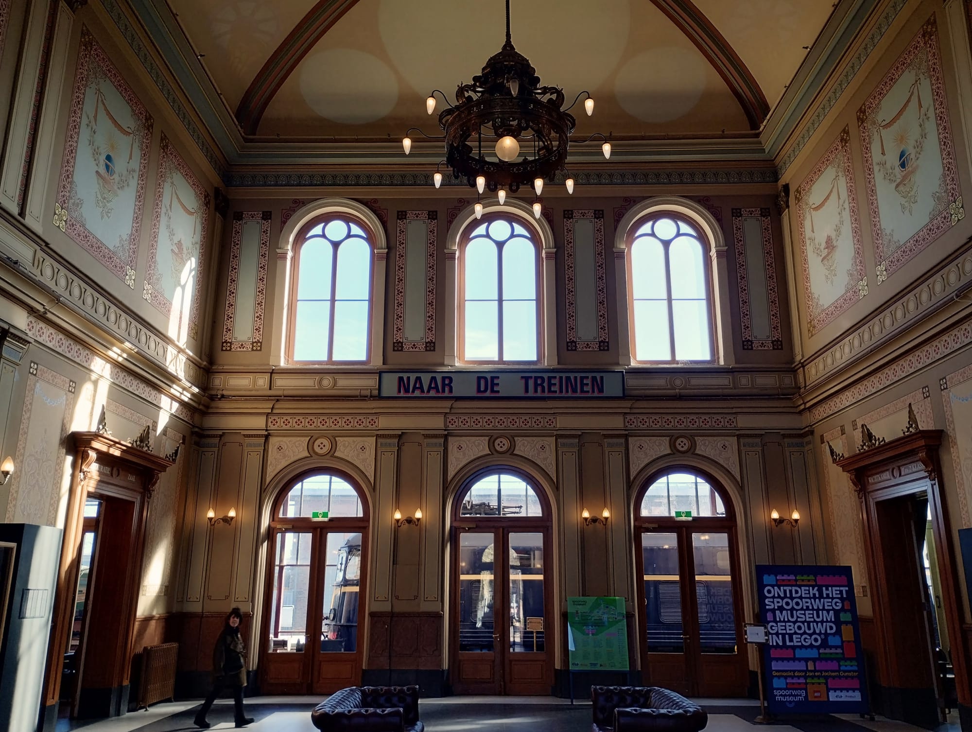 Interior of a train station hall with ornate architecture and a large chandelier, featuring arched windows with the text "NAAR DE TREINEN" (To the Trains) above them, and a person walking on the left side, along with a poster advertising a LEGO museum on the right.