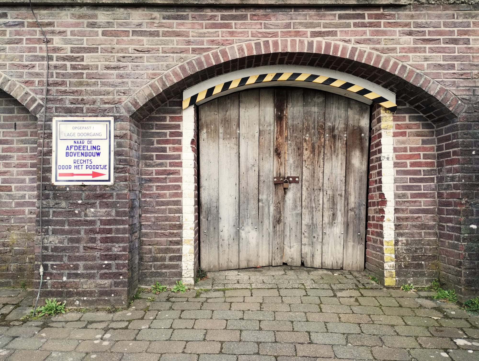 Weathered wooden door with a lock, under a brick archway with a black and yellow striped warning, set in a brick wall with a Dutch language sign indicating a low doorway and rightward access to the upper level of the building.