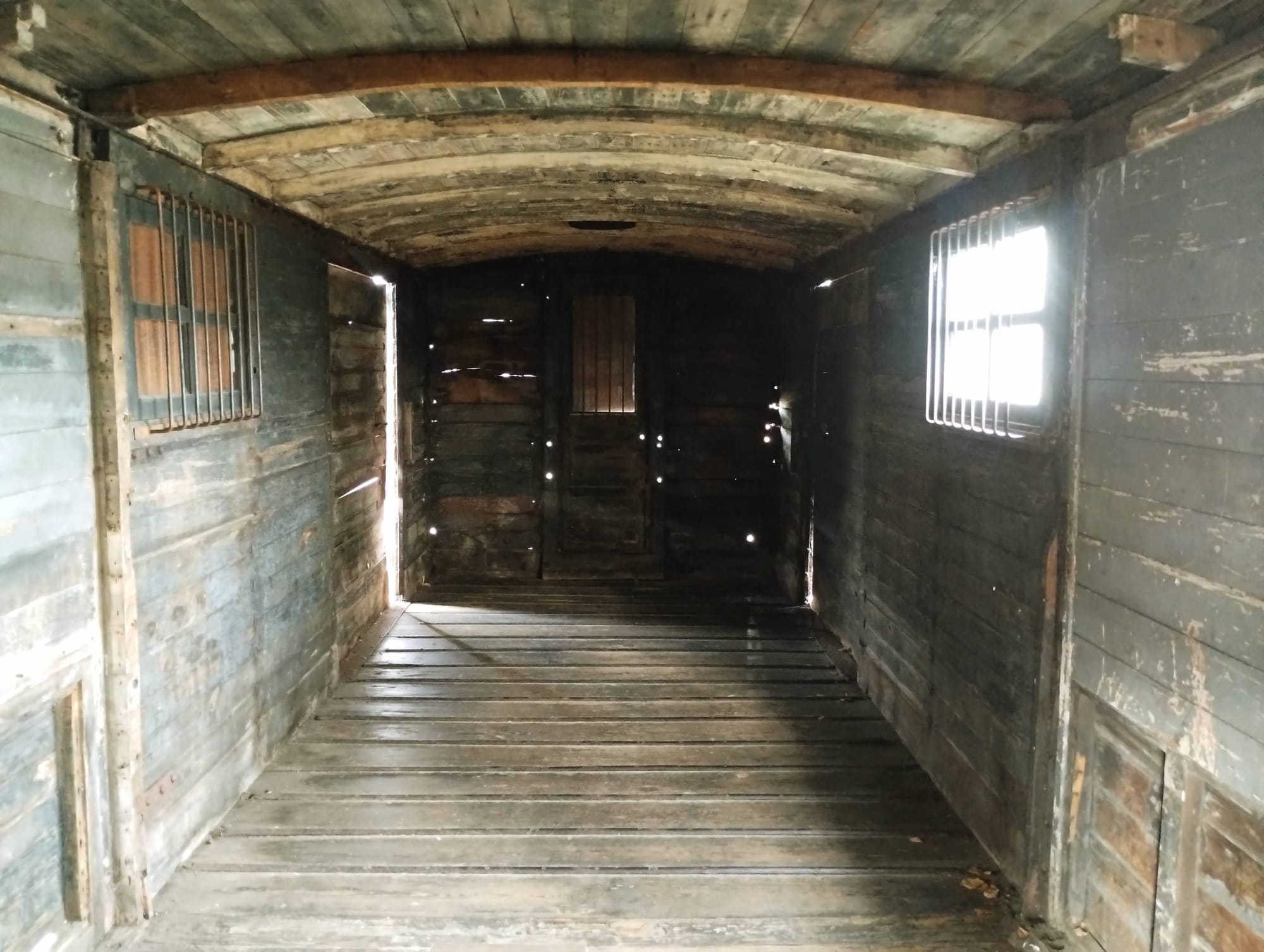 Interior view of an old, weathered wooden train car, with the arched ceiling and walls composed of aged, grey horizontal planks. The floor is also constructed of wooden planks, with a long, linear perspective towards the dark rear of the car. Light streams in from barred windows on either side, illuminating dust motes and creating a contrast of light and shadow within the car.