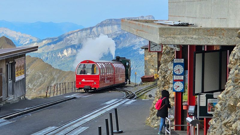 A red cogwheel train with a green engine steams along a mountain track at a station overlooking a panoramic mountain range.