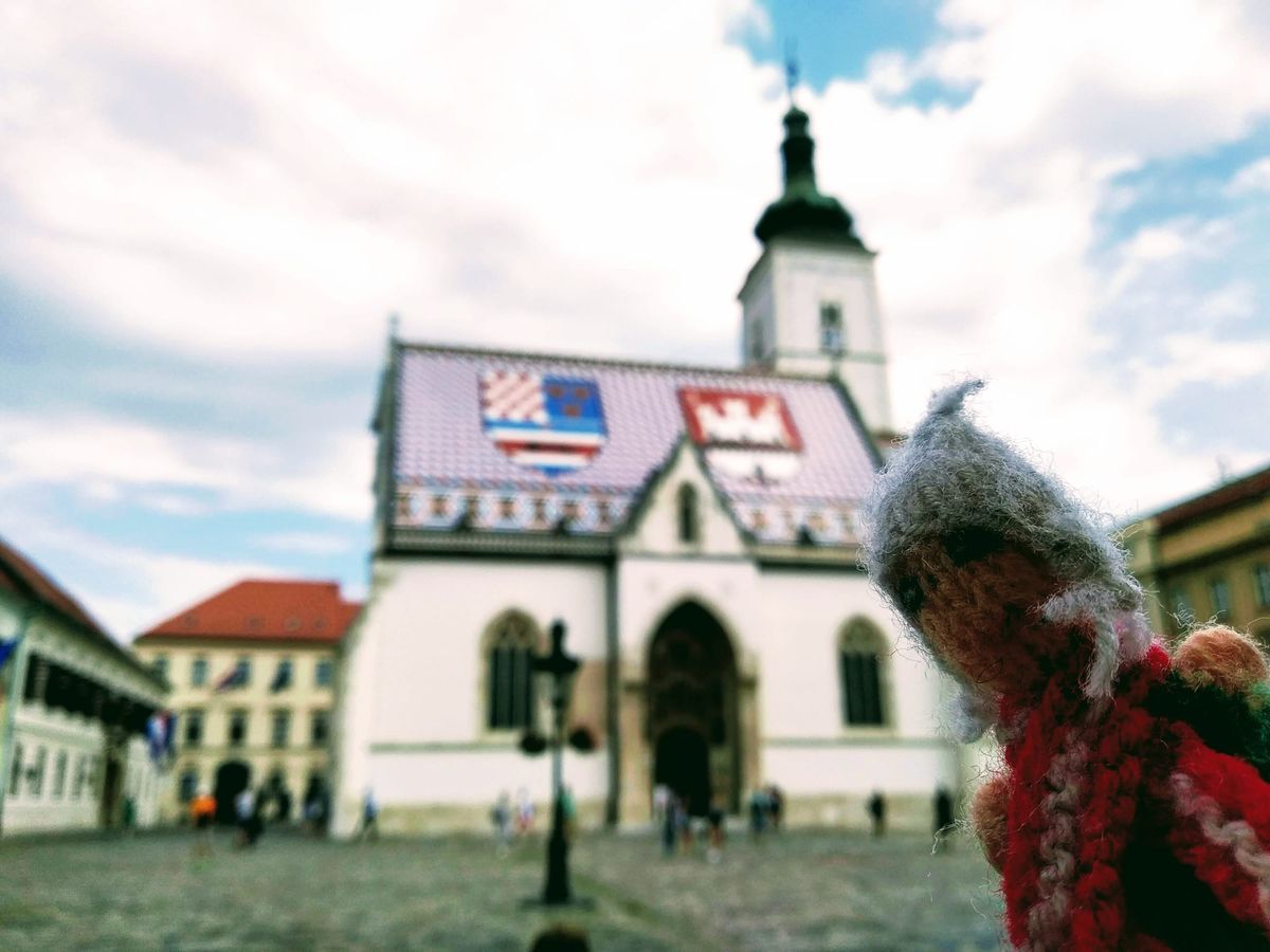 Finger puppet looking pious in front St. Mark's Church Croatia, featuring its distinctive tiled under a cloudy sky, with tourists in the square and surrounding buildings.
