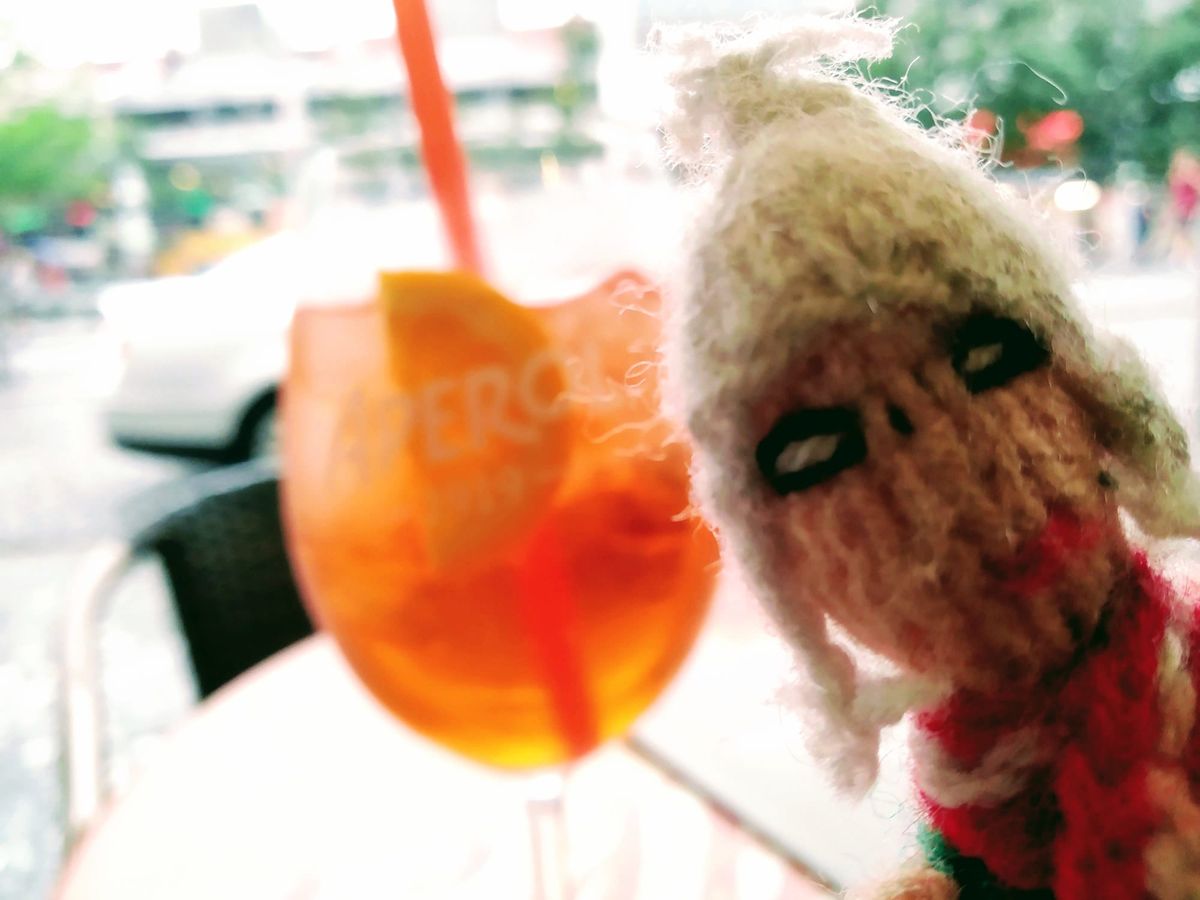 A dipsomaniac finger puppet next to a soon-to-be-empty glass of Aperol spritz, with a blurred street scene visible through a window in the background.
