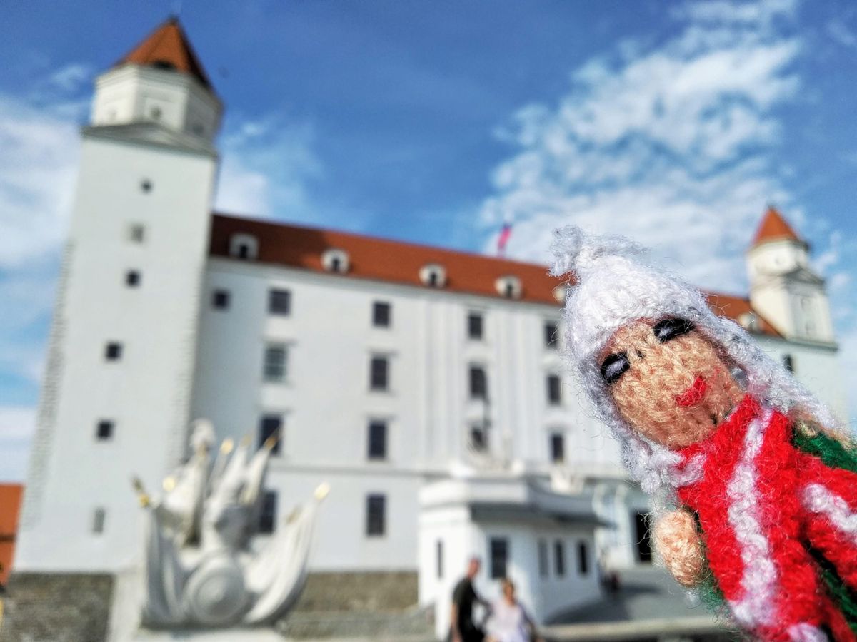 An excessively perky  finger puppet in a white hat and red and white striped jacket in front of the white castle in Bratislava.