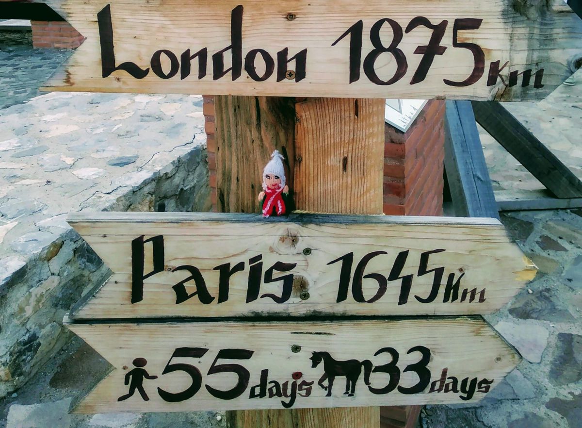 Finger puppet examines a wooden signpost pointing to London (1875 km) and Paris (1645 km). Travel times on foot (55 days) and by horse (33 days).
