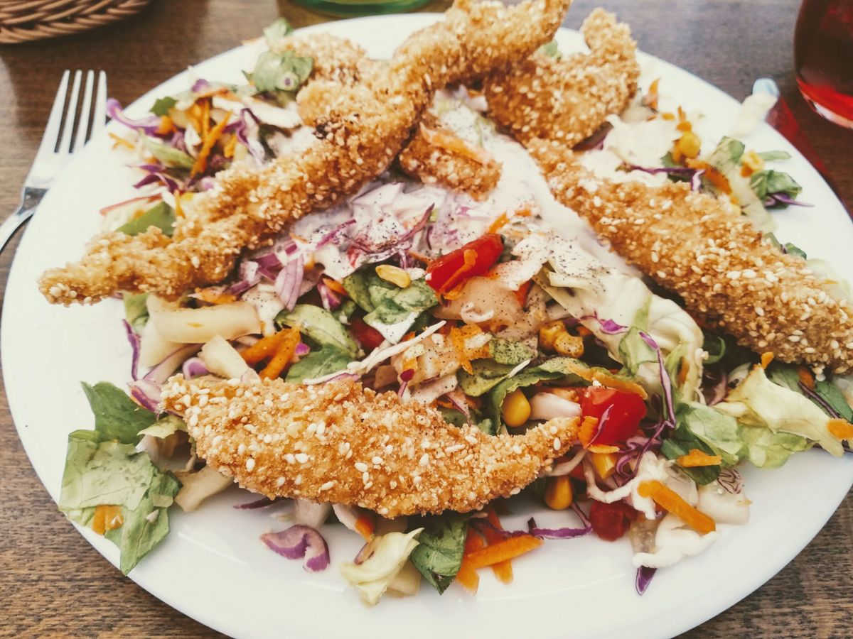 A white plate holding a salad with fried sesame-crusted chicken tenders on top, and mixed greens, shredded red cabbage, carrots, and tomatoes.