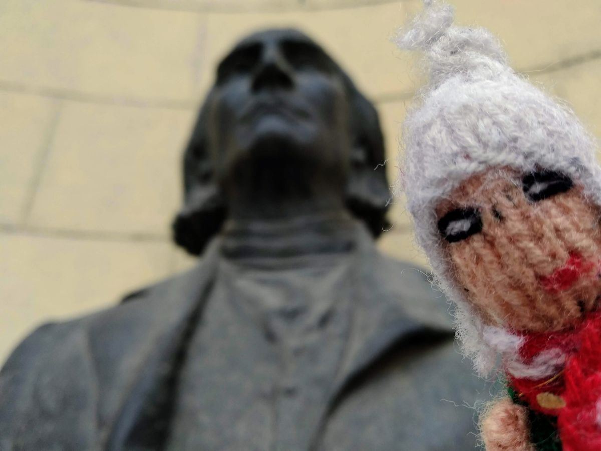 A finger puppet wearing a white hat and red scarf is positioned in front of a blurred bronze statue of a man, likely a historical figure.